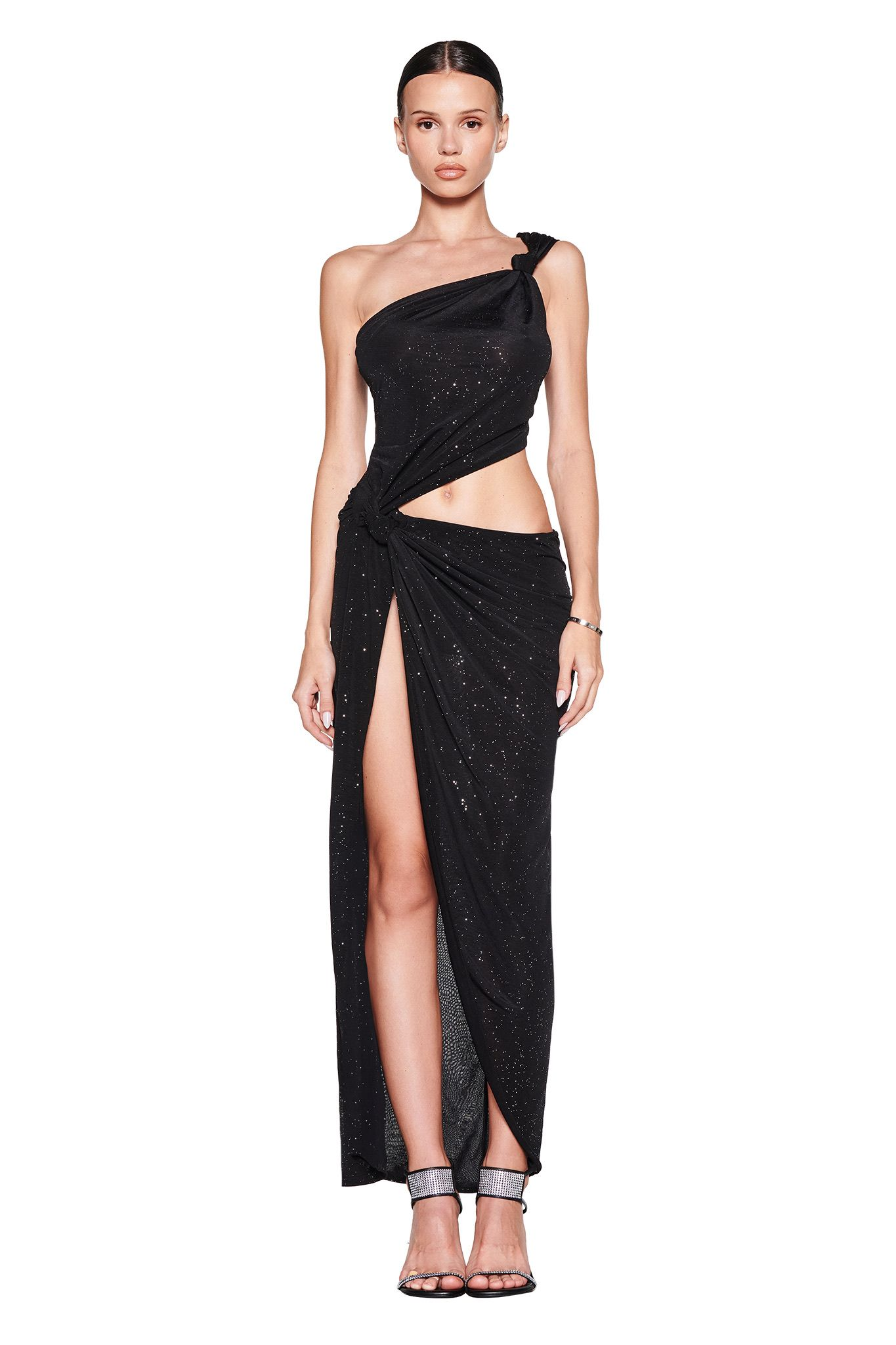 A woman stands facing forward in the I.AM.GIA ALIX MAXI DRESS—an asymmetrical black, sparkly, one-shoulder gown featuring a high slit and waist cutout. She has slicked-back hair, strappy heels, and minimal jewelry against a plain white background.