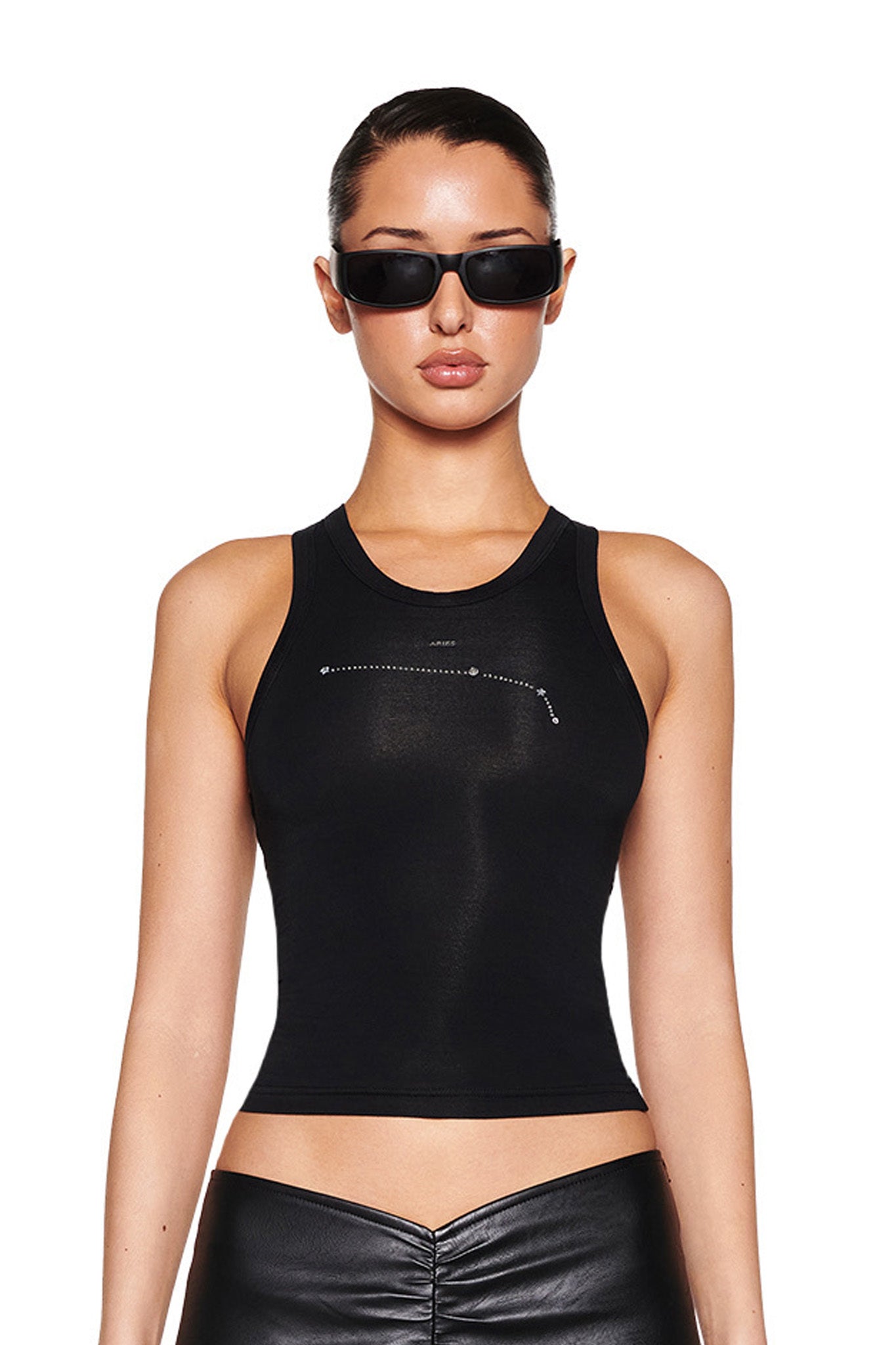 A woman models the I.AM.GIA ARIES ZODIAC TANK - BLACK with sleek leather pants and chic black sunglasses. Against a plain white backdrop, the tank features a subtle Aries constellation graphic as she gazes at the camera with a neutral expression.