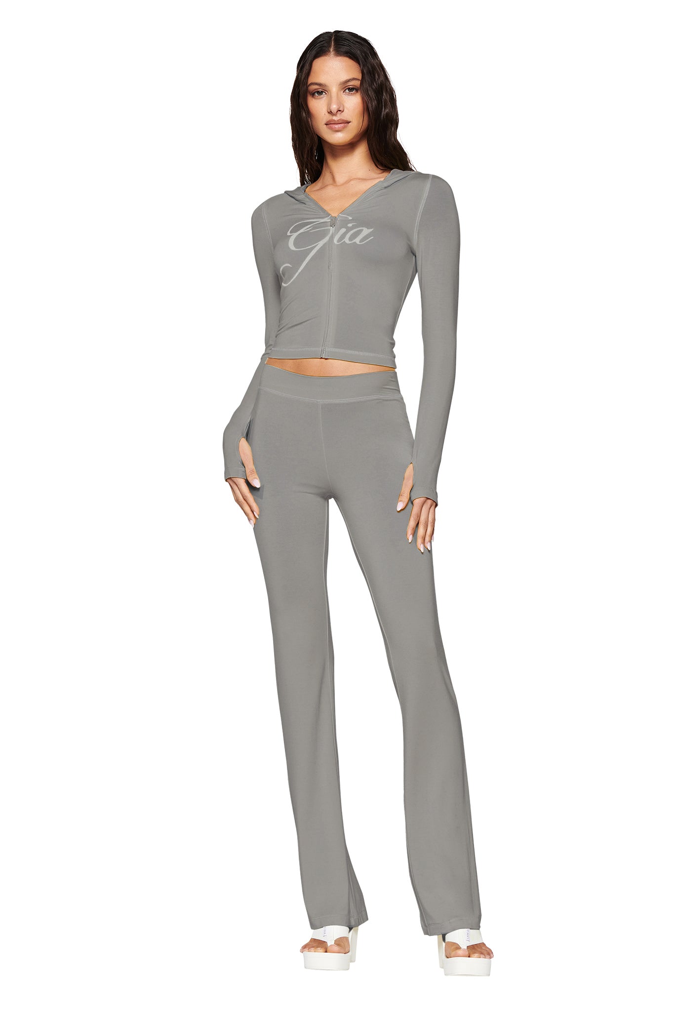 A woman showcases a bodycon look, wearing the I.AM.GIA "Blare" hoodie in sage with matching high-waisted pants and white platform sandals, all against a plain white background.