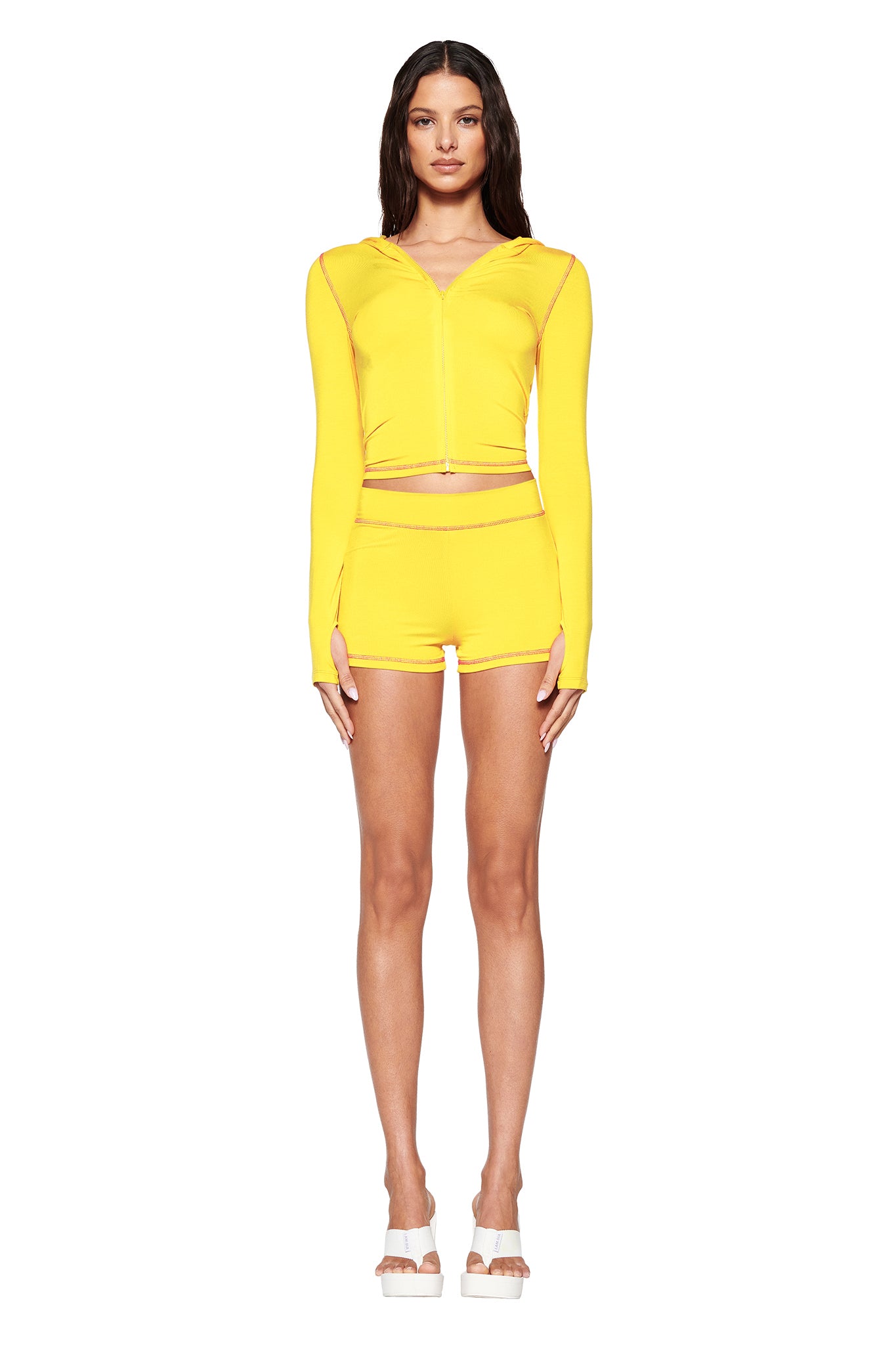 Dressed in I.AM.GIA's BLARE HOODIE - LEMON and matching shorts, a person showcases a sleek bodycon look against a plain white backdrop, complemented by white platform sandals.