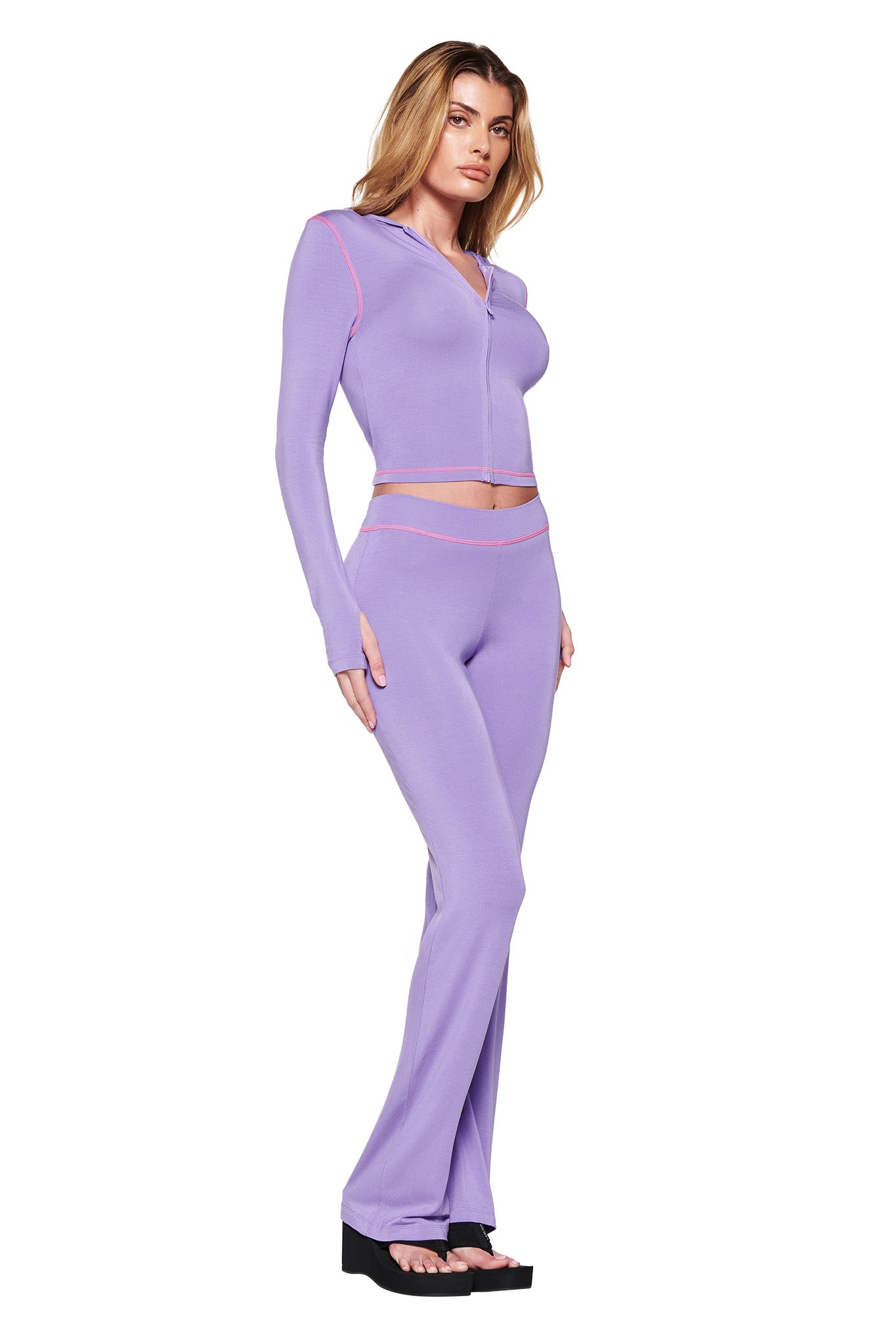 A woman poses in a chic lilac ensemble from I.AM.GIA, featuring the BLARE HOODIE - PURPLE with pink accents and flared pants. She finishes the look with black platform sandals against a plain white backdrop.