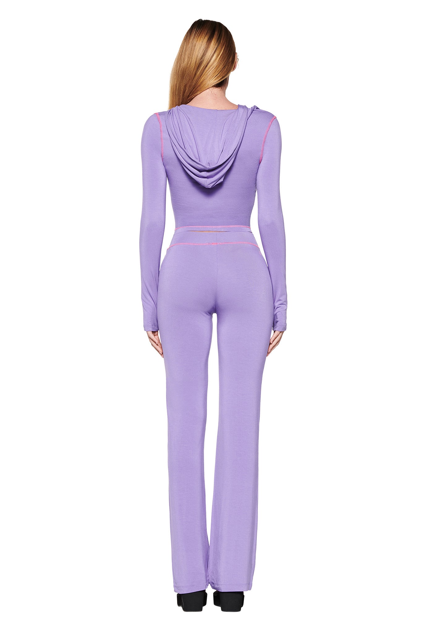 Facing away, a model showcases the BLARE TRACKPANT - PURPLE by I.AM.GIA. The fitted jumpsuit features long sleeves, flared legs, and a hood in a stretchy bamboo fabric. With straight hair flowing down, black shoes complete the look against the white backdrop.