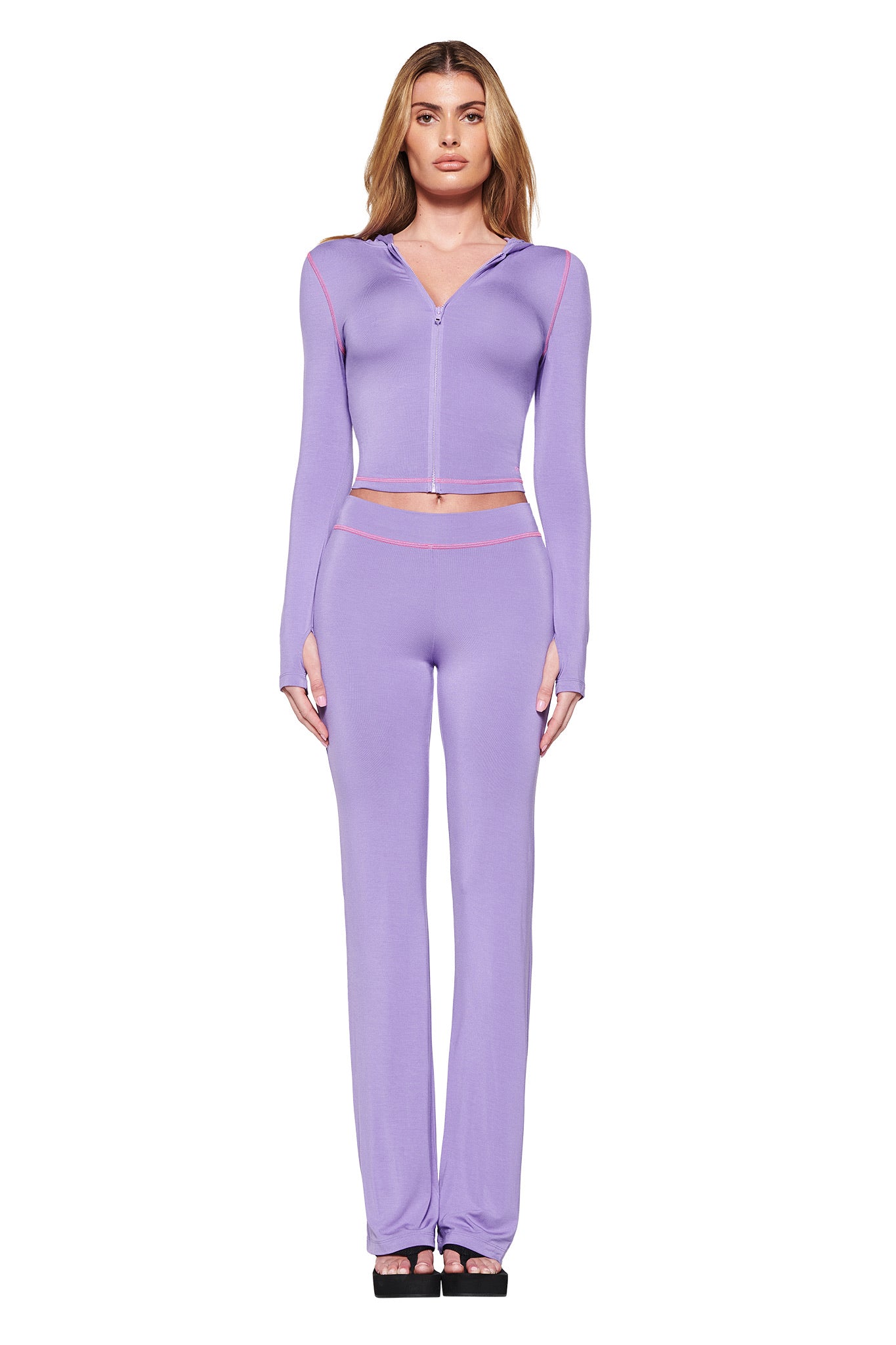 A woman wears the BLARE HOODIE in purple by I.AM.GIA, a bodycon zip-up top with pink seam details, paired with high-waisted pants. She stands against a white background and completes the look with black platform sandals.