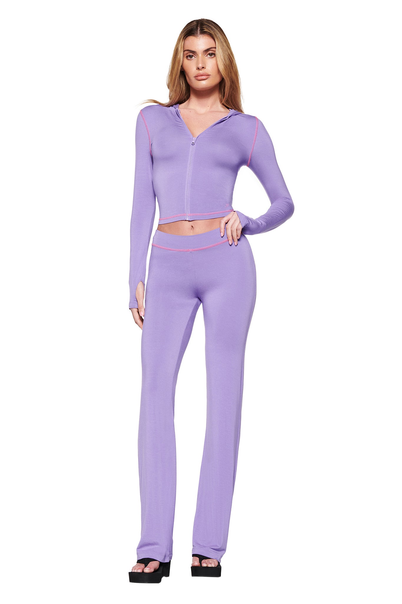 A person models an I.AM.GIA purple outfit against a white background, featuring a light purple, long-sleeve crop top with zip-up design and matching BLARE TRACKPANT high-waisted flared pants with coral trim, complemented by black platform sandals.