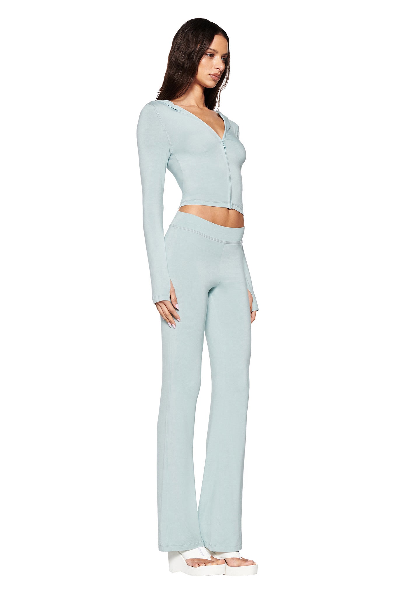 A person wearing an I.AM.GIA Seafoam Blare Trackpant, low-rise with flared legs, styled with long hair and white platform sandals, poses against a white background.