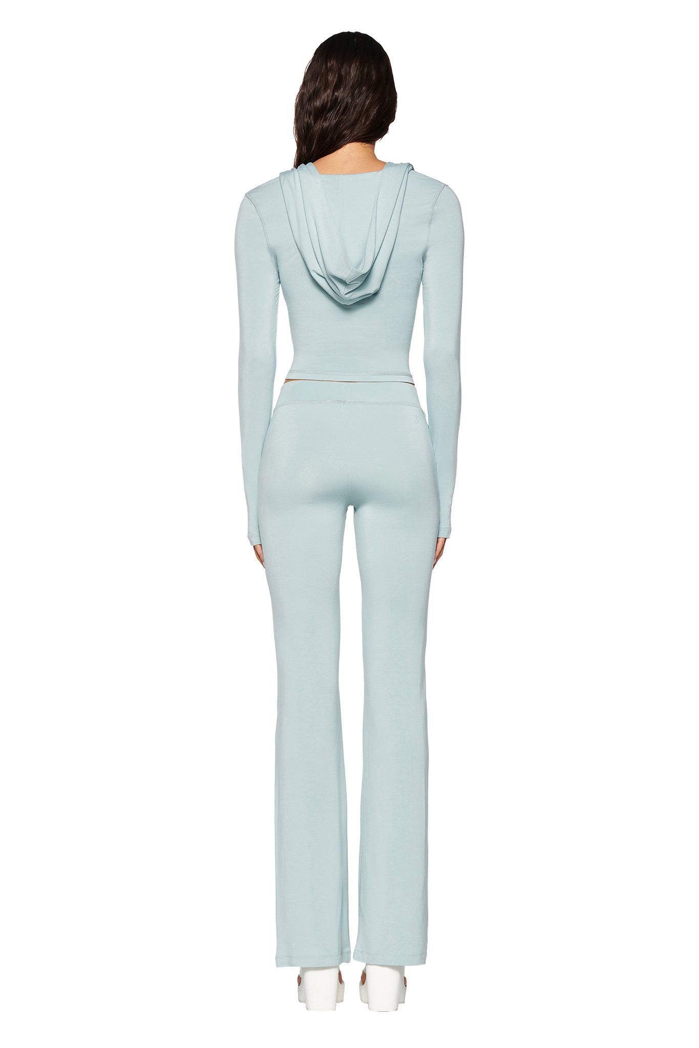 Facing away, they wear a light blue hooded top with I.AM.GIA's BLARE TRACKPANT - SEAFOAM, featuring a low-rise waist. Their long dark hair contrasts the minimal white backdrop, and they complete the look with white heeled shoes. The bamboo and jersey fabric adds elegance.