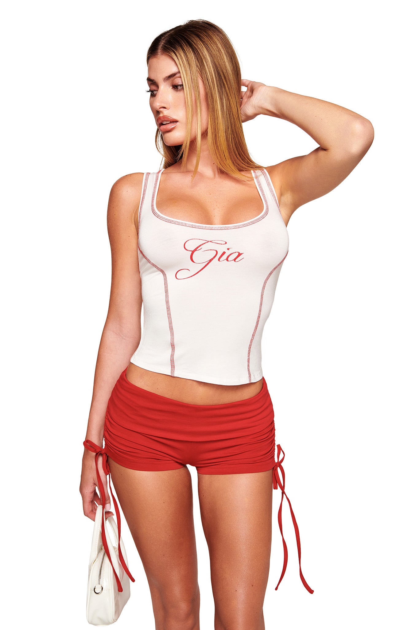 A woman with long blonde hair wears the I.AM.GIA BLARE TANK in white with "Gia" on it, paired with red ruched side-tie shorts. She holds a small white handbag and looks to the side against a white background.