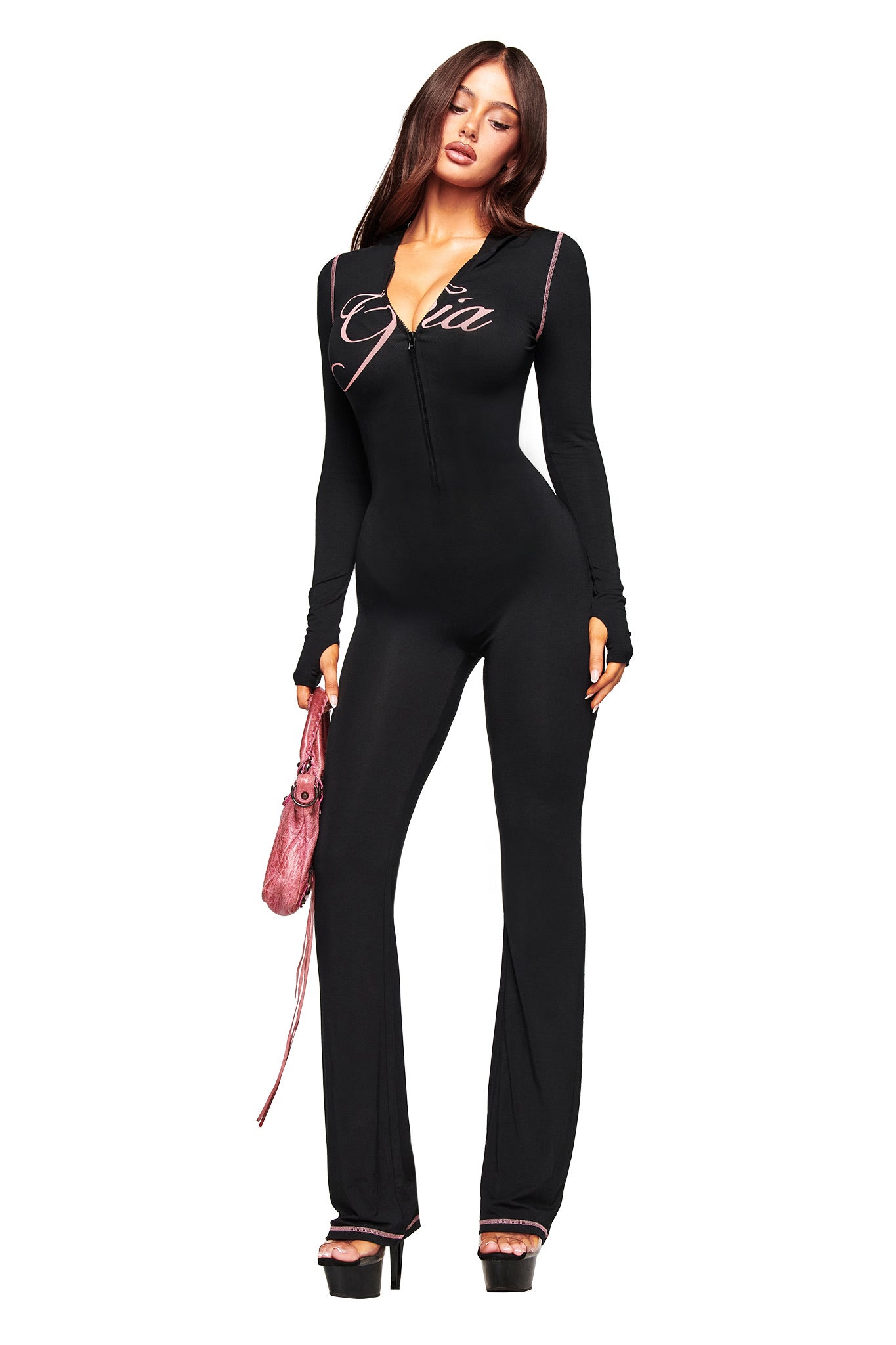 A woman poses against a white background in the I.AM.GIA BLARE JUMPSUIT - BLACK, a fitted style with flared legs and "Gia" on the chest. She accessorizes with a pink purse and black platform heels.