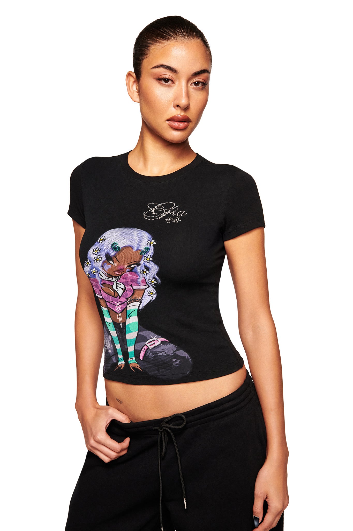 A woman with slicked-back hair models the FAFI T-SHIRT - BLACK by I.AM.GIA, featuring colorful cartoon artwork, paired with black pants and posing against a white background with one hand in her pocket.