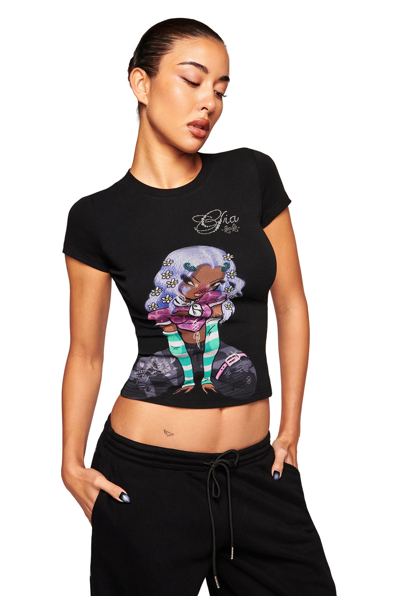 A woman with slicked-back hair models the I.AM.GIA FAFI T-SHIRT - BLACK, showcasing a vibrant cartoon character with striped green socks and purple hair standing between two pumpkins. She poses with one hand in her pocket against a white background.