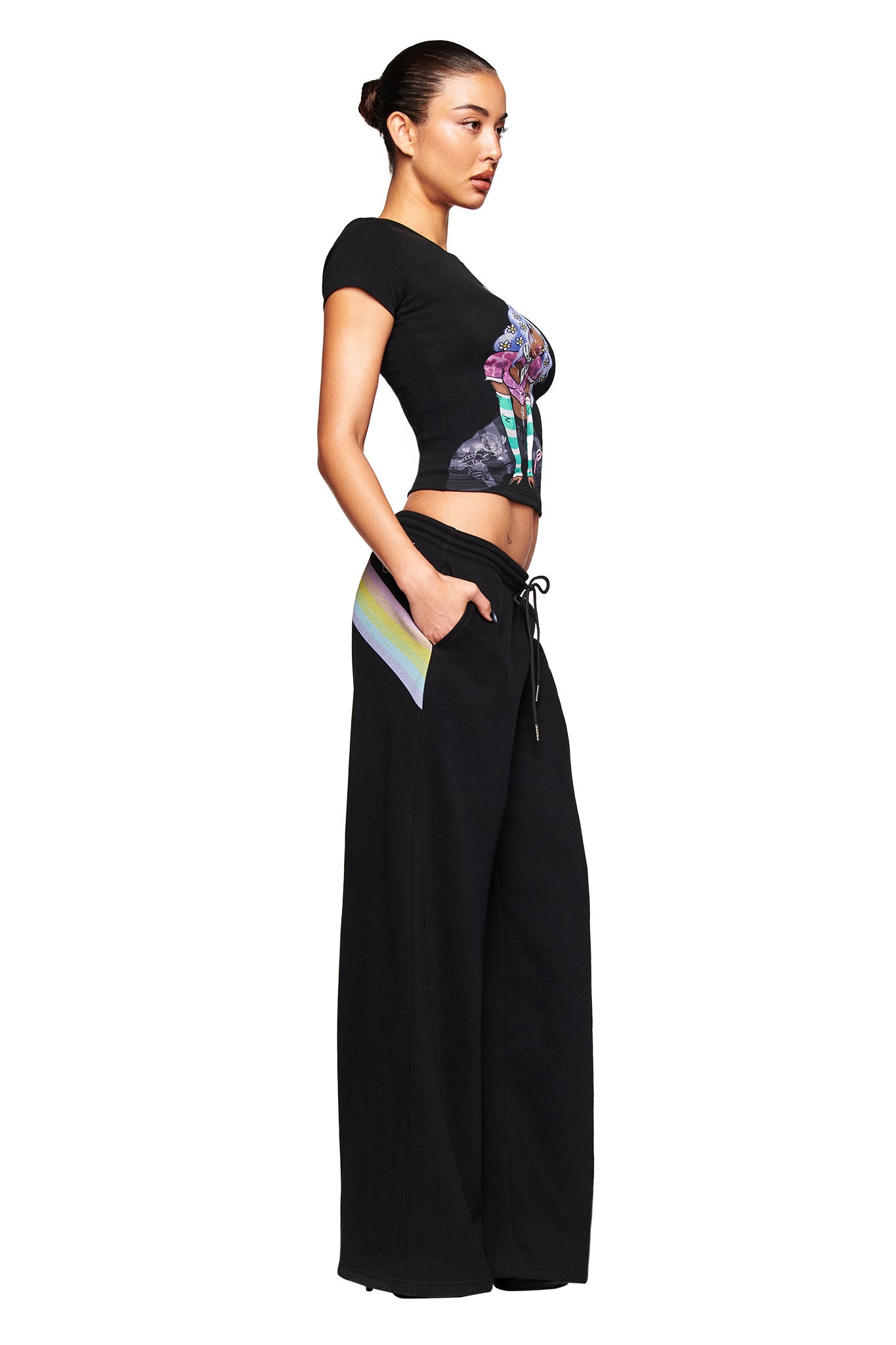 A woman stands in profile against a white background wearing the I.AM.GIA FAFI T-SHIRT - BLACK with a colorful graphic, paired with loose black wide-leg pants and her hands in the pockets.