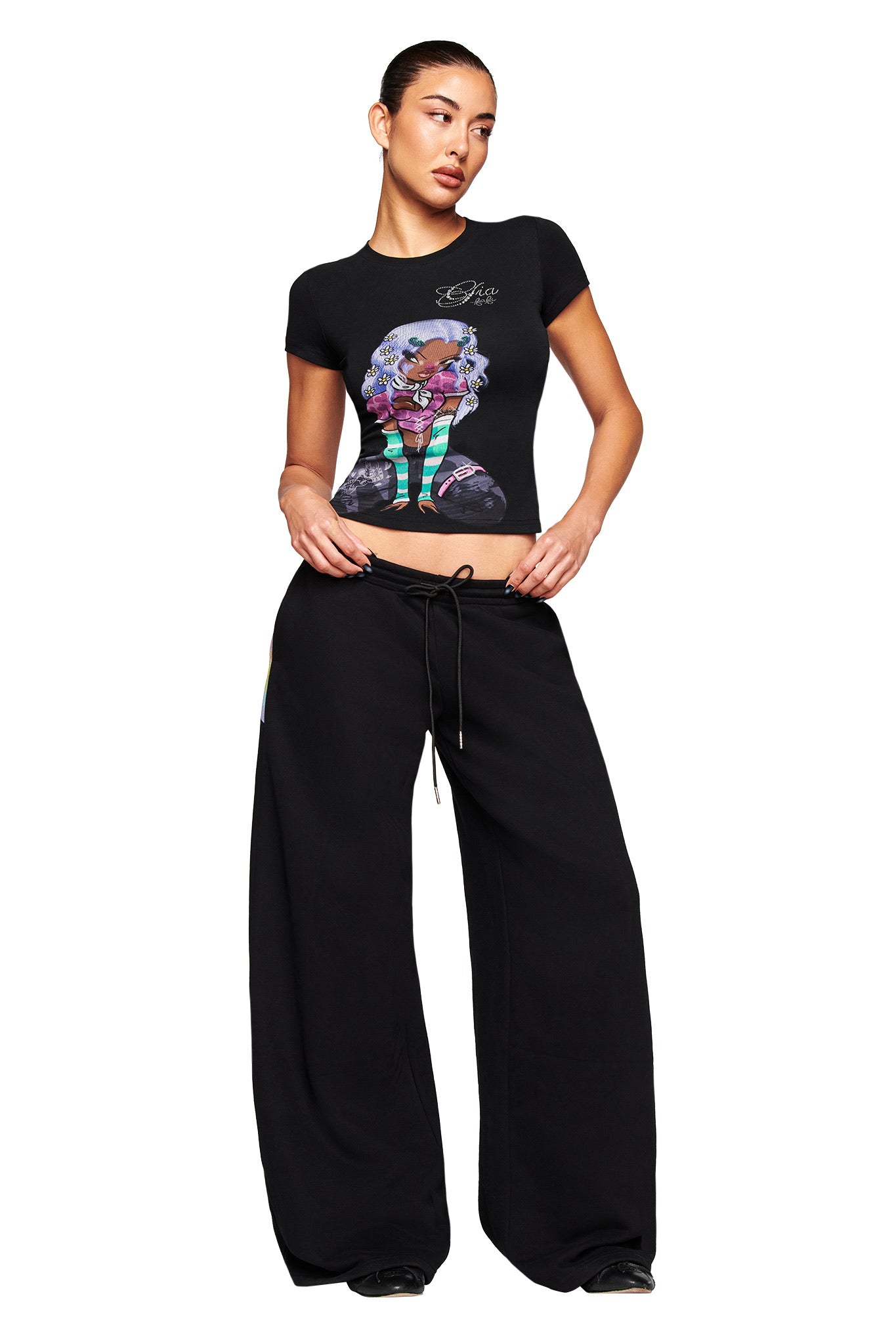 A woman stands against a white background wearing the I.AM.GIA FAFI T-SHIRT in black, featuring a colorful animated graphic, paired with loose black drawstring pants. She poses confidently with her hands on her hips, gazing to the side.