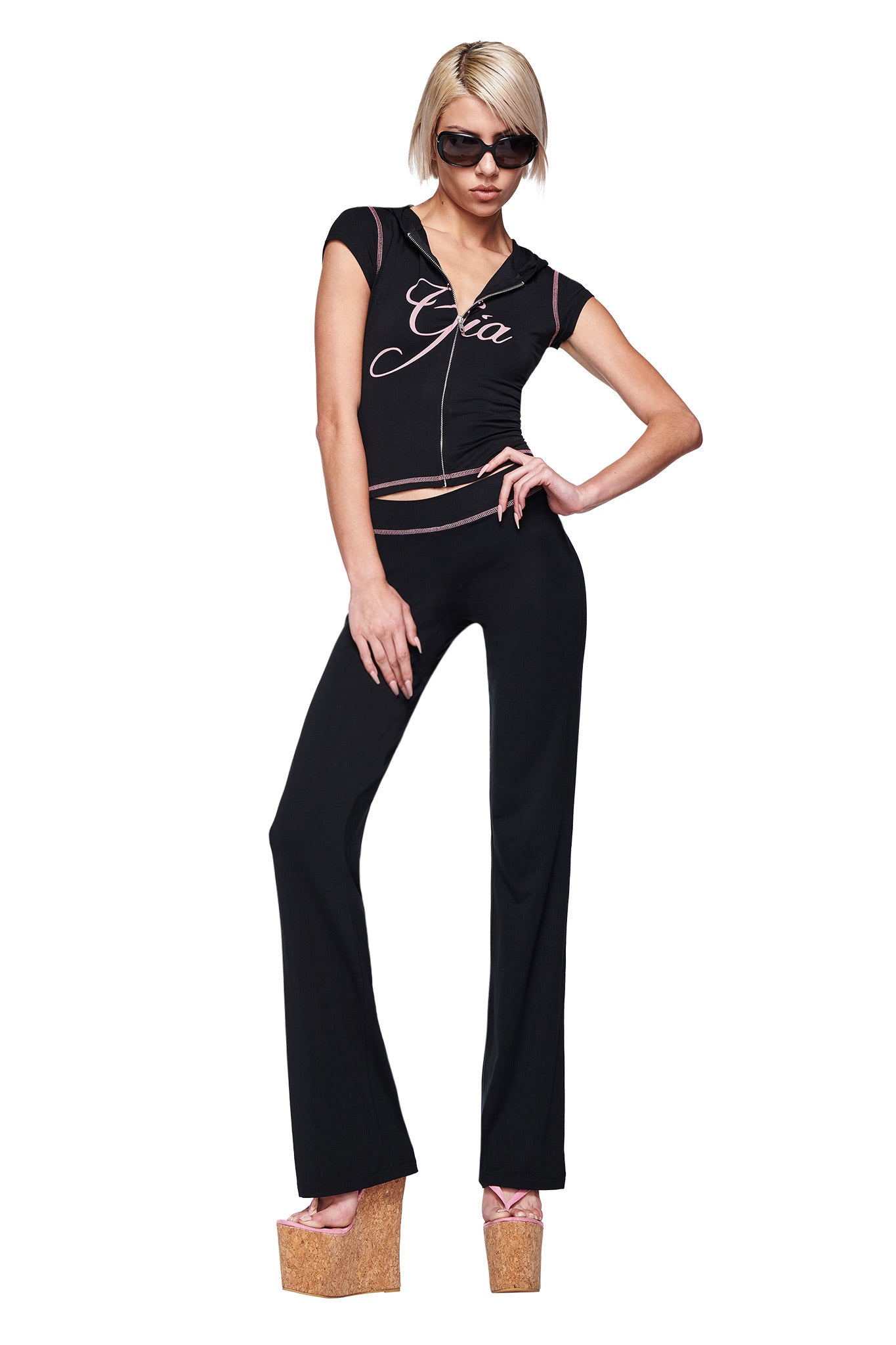 A woman wearing black sunglasses, the I.AM.GIA BLARE CAP SLEEVE HOODIE in black, matching black pants, and high cork platform sandals poses confidently with one hand on her hip.