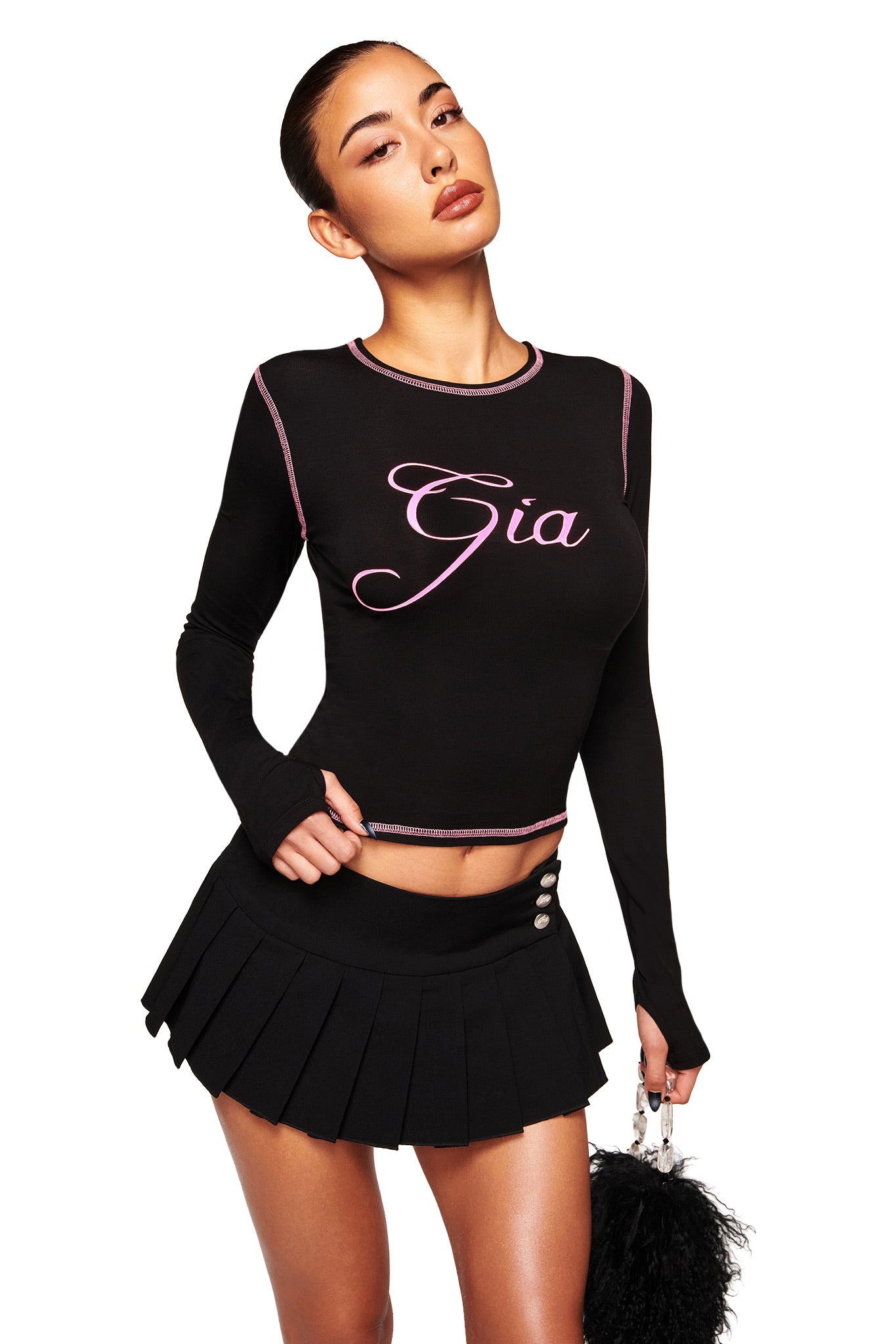 A woman stands confidently in a black I.AM.GIA BLARE LONG SLEEVE TOP featuring "Gia" in pink script, paired with a black pleated mini skirt and holding a black fluffy handbag, set against a plain white background.
