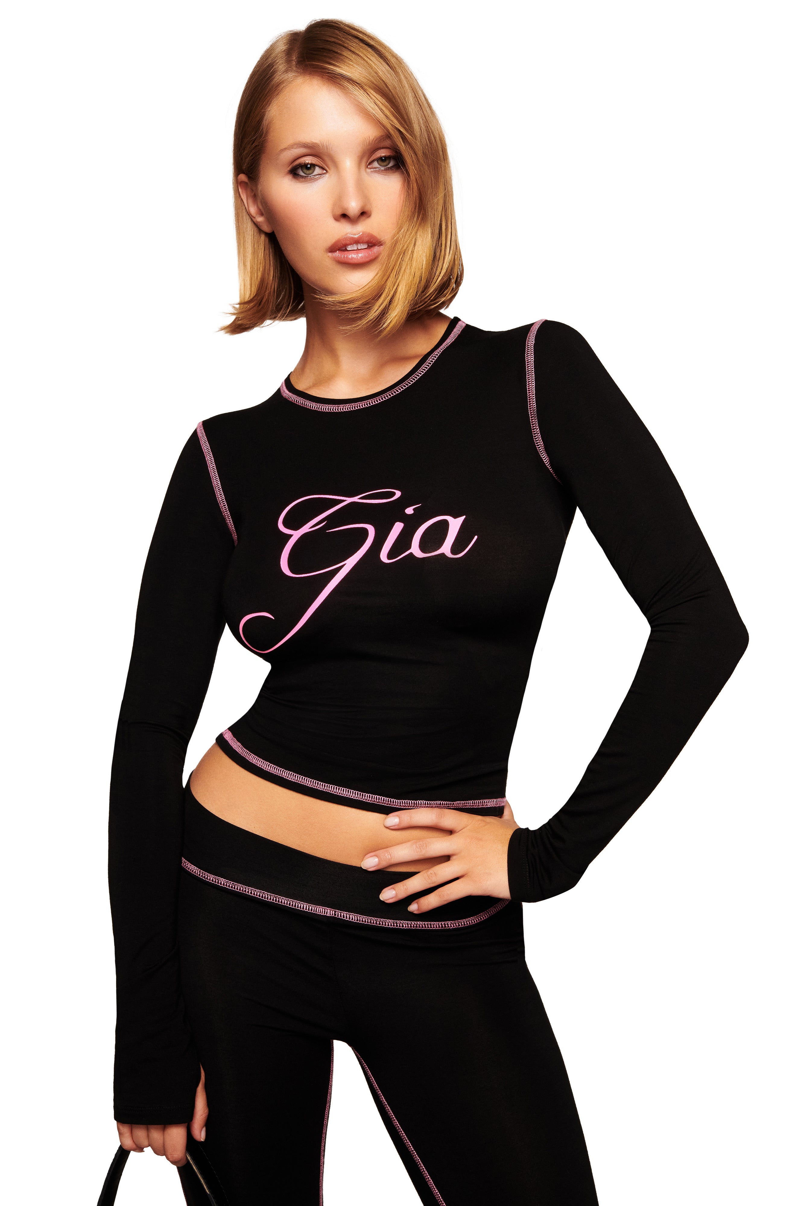 A woman with shoulder-length blonde hair wears the I.AM.GIA BLARE LONG SLEEVE TOP in black, featuring pink trim and "Gia" script. She pairs it with matching pants, posing confidently with one hand on her hip.
