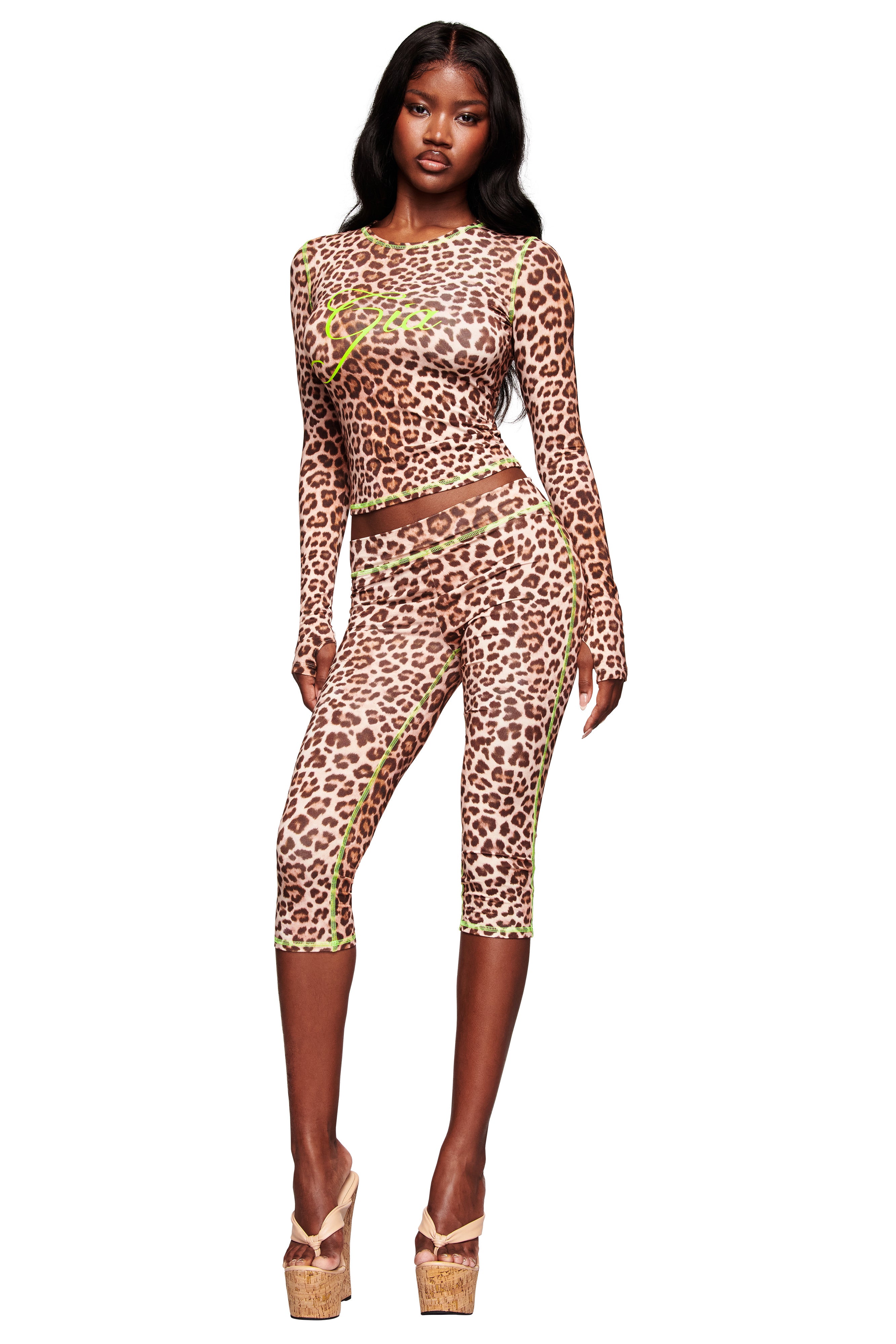 A woman models the I.AM.GIA BLARE CAPRI - LEOPARD: a fitted, long-sleeve leopard print top with matching capri pants featuring neon green seams and tan platform heels, set against a white background.
