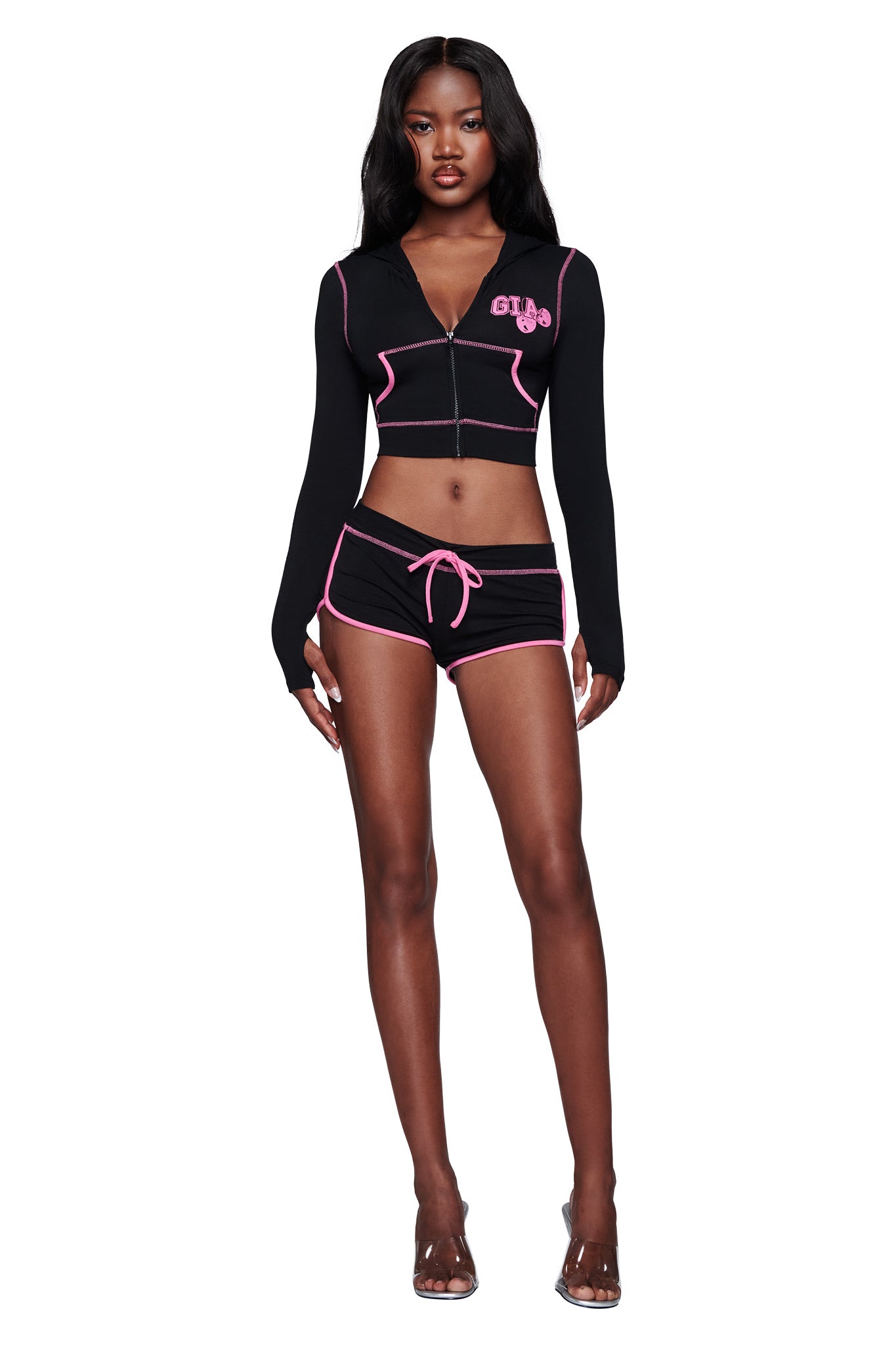 A woman stands wearing the I.AM.GIA BLARE CROPPED HOODIE in black, paired with matching black shorts featuring pink trim and a drawstring, and clear heels, against a white background.