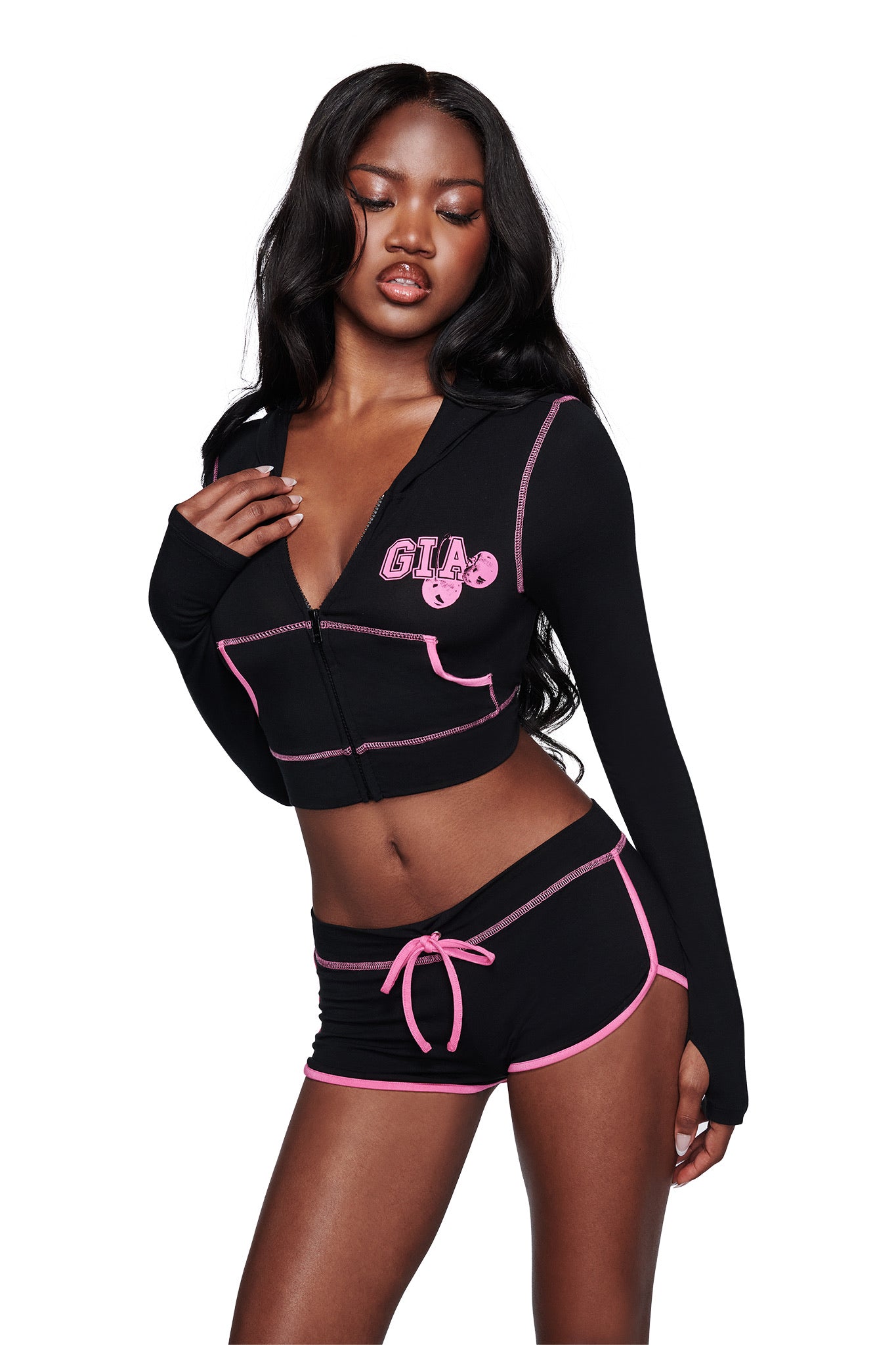 A woman poses against a white background wearing the I.AM.GIA BLARE CROPPED HOODIE in black, featuring "GIA" in pink letters, paired with matching black shorts detailed with pink trim and a front tie.