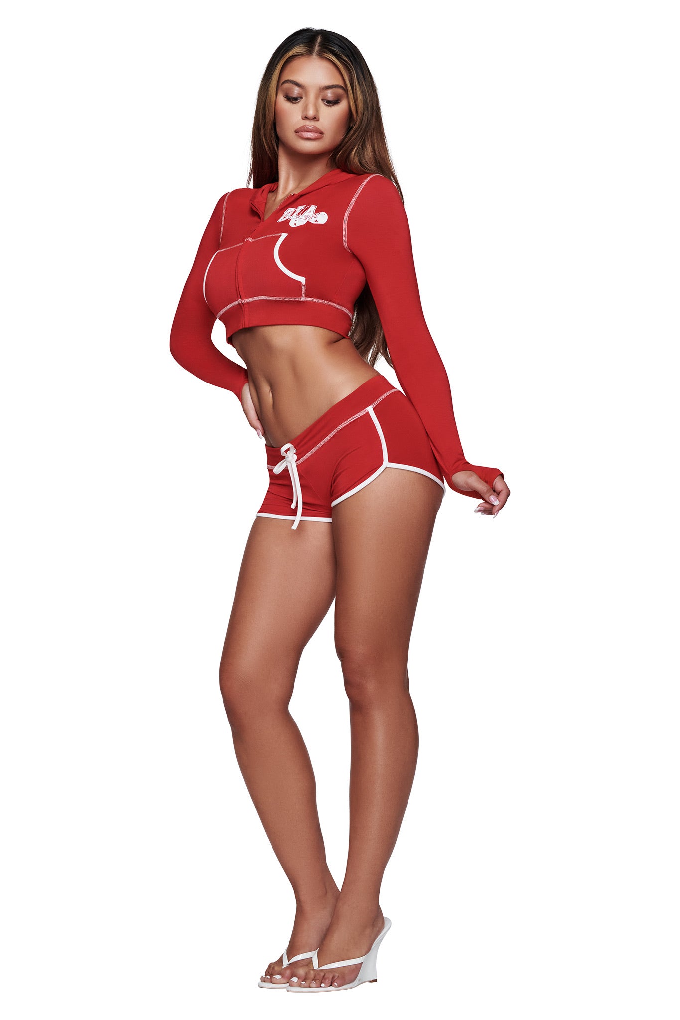 A woman poses against a white background wearing the I.AM.GIA BLARE CROPPED HOODIE in red, paired with matching red shorts featuring white trim and clear high-heeled shoes.