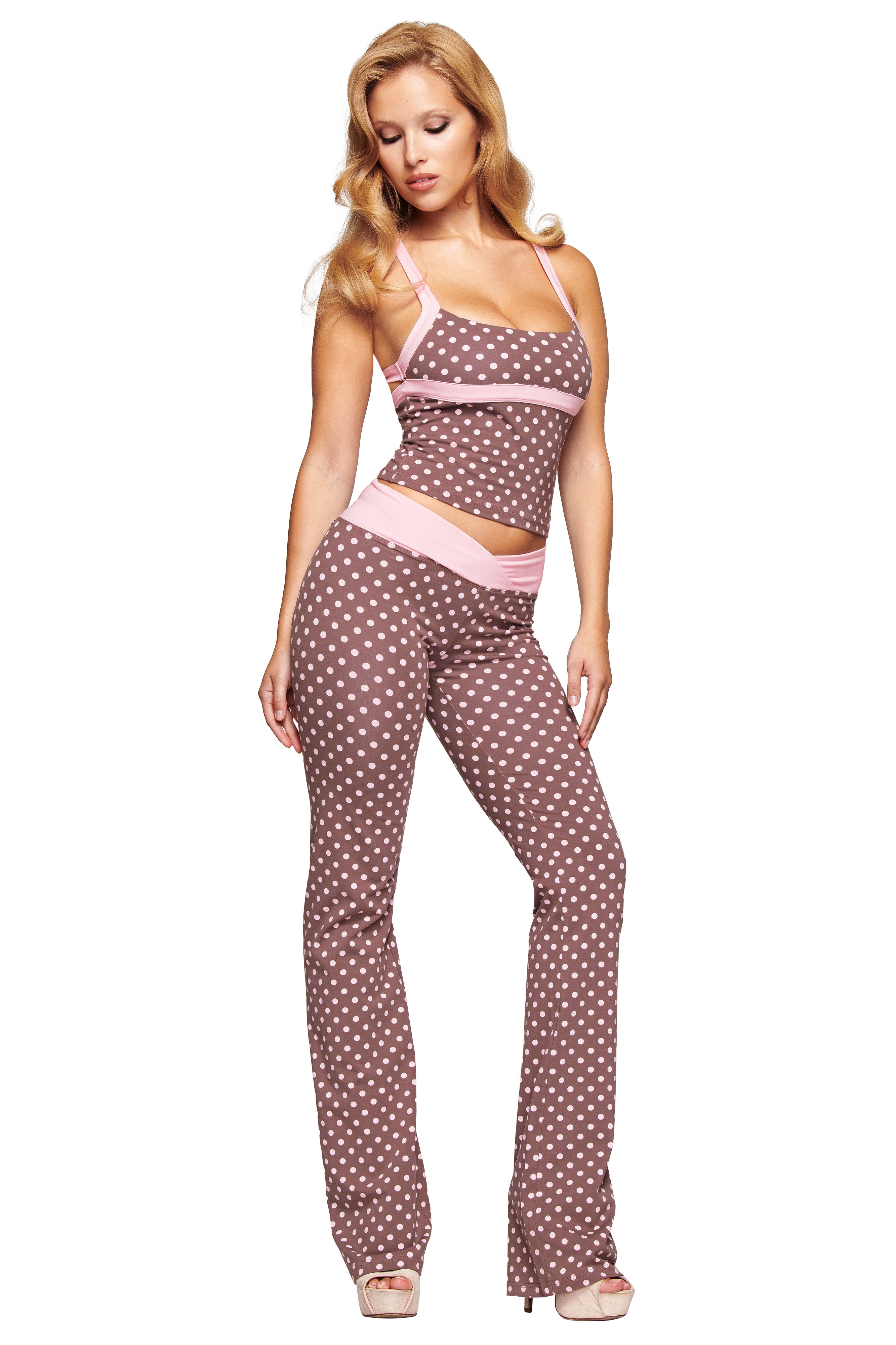 A woman wearing I.AM.GIA's Blare Crossover Pant in Pink Chocolate, featuring a brown sleeveless polka dot print and flared legs with light pink trim, poses in high heels against a white background.