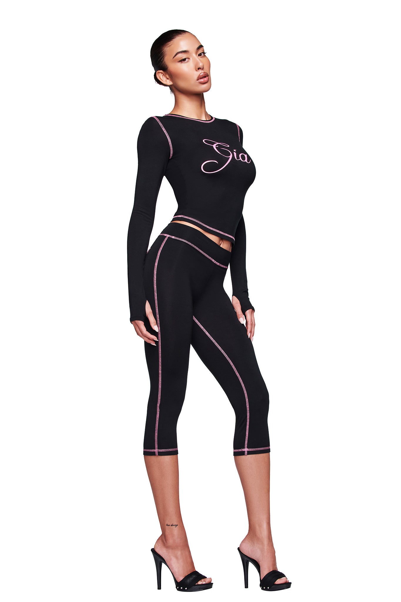 A woman models the I.AM.GIA BLARE CAPRI in black with pink stitching and a bodycon fit, paired with a matching black crop top that reads "Gia" and black high-heeled sandals, posing against a white background.