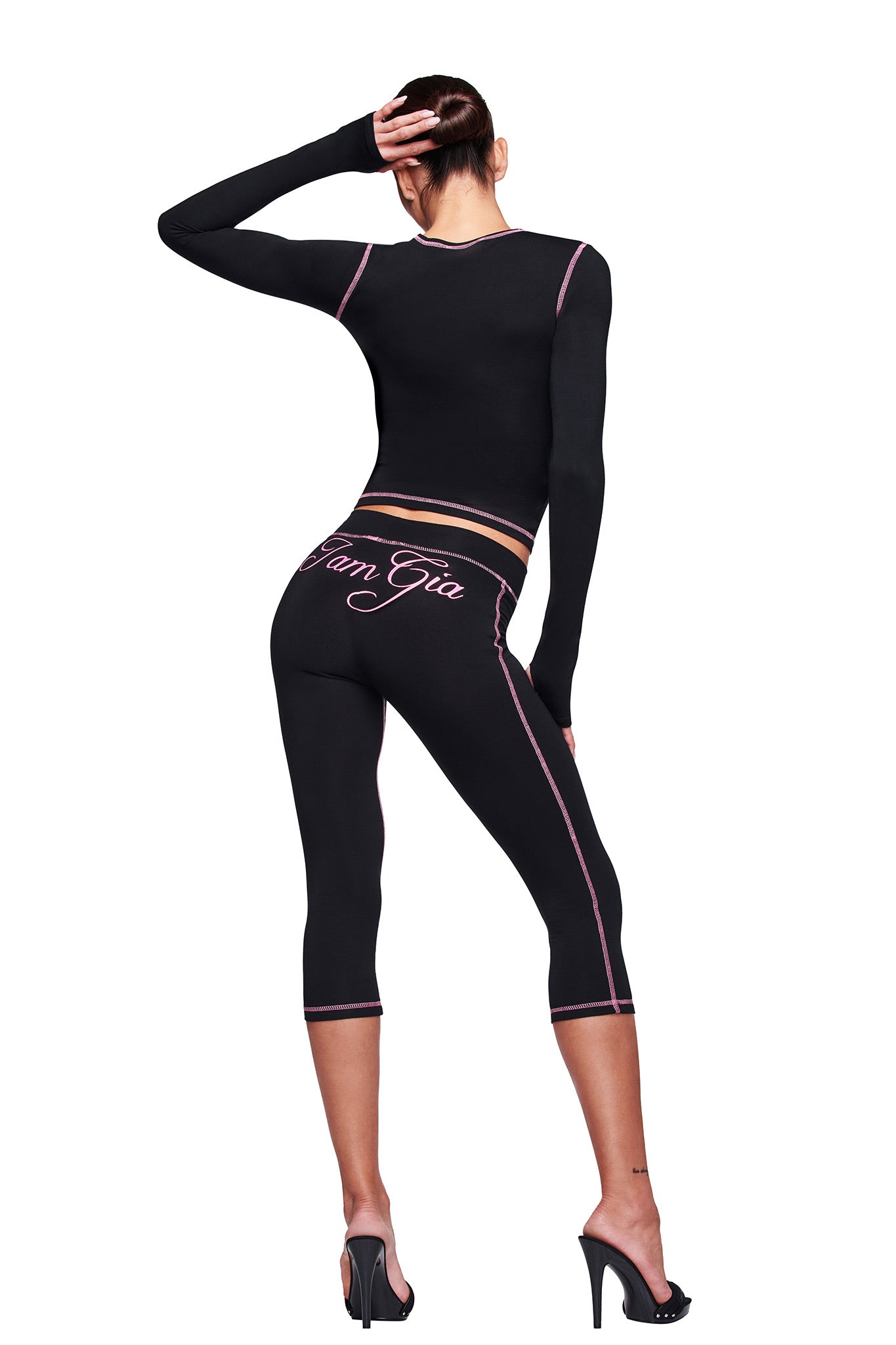 A woman stands with her back to the camera, one hand on her head, wearing the I.AM.GIA BLARE LONG SLEEVE TOP - BLACK paired with fitted pink-trim activewear and black high heels.