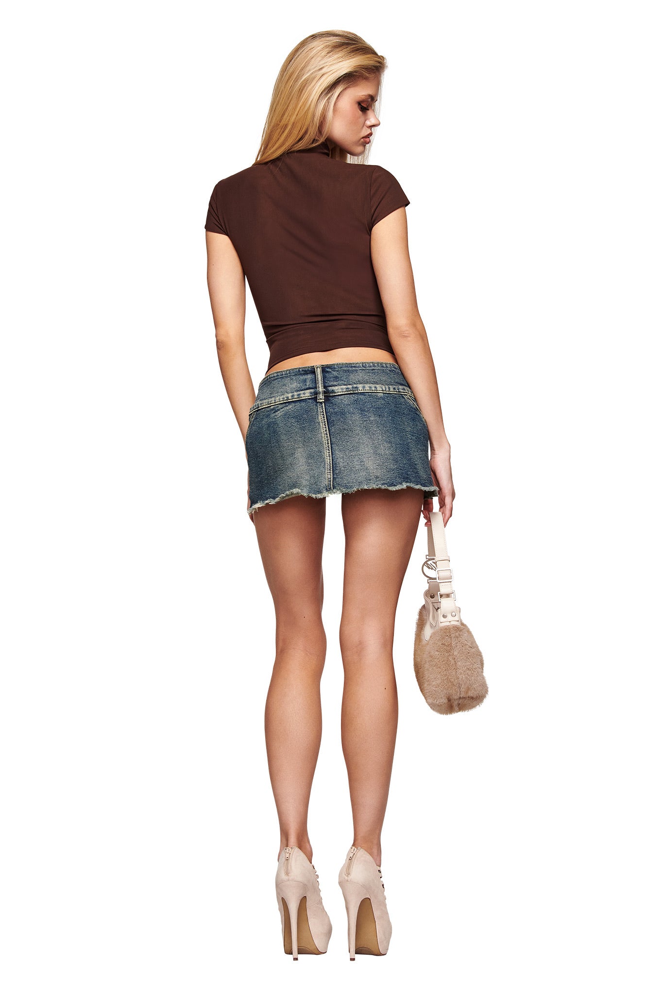 A woman with long blonde hair, standing against a white background, wears the I.AM.GIA ISELA TOP in brown, paired with a short denim skirt, beige high heels, and holds a beige furry handbag.
