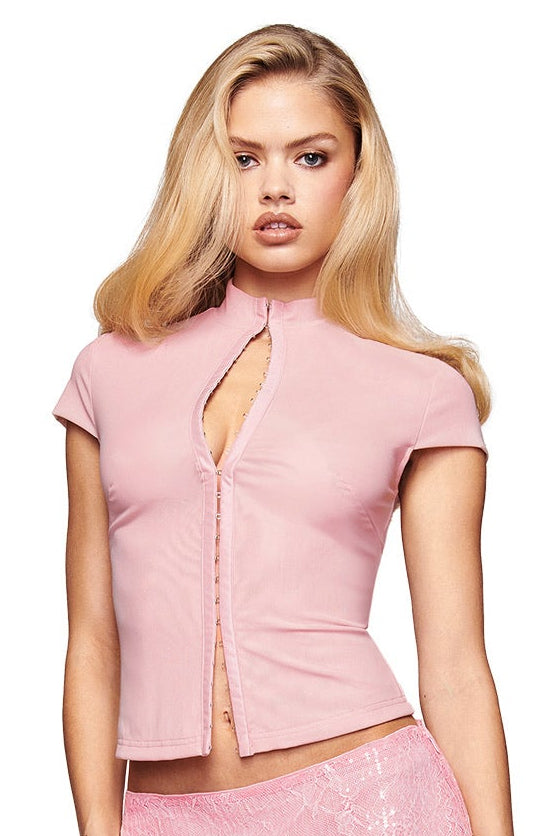 A woman in the I.AM.GIA ISELA TOP - BLUSH, paired with a pink sequin mini skirt, platform heels with ribbon laces, and a small pink purse, stands against a white background.