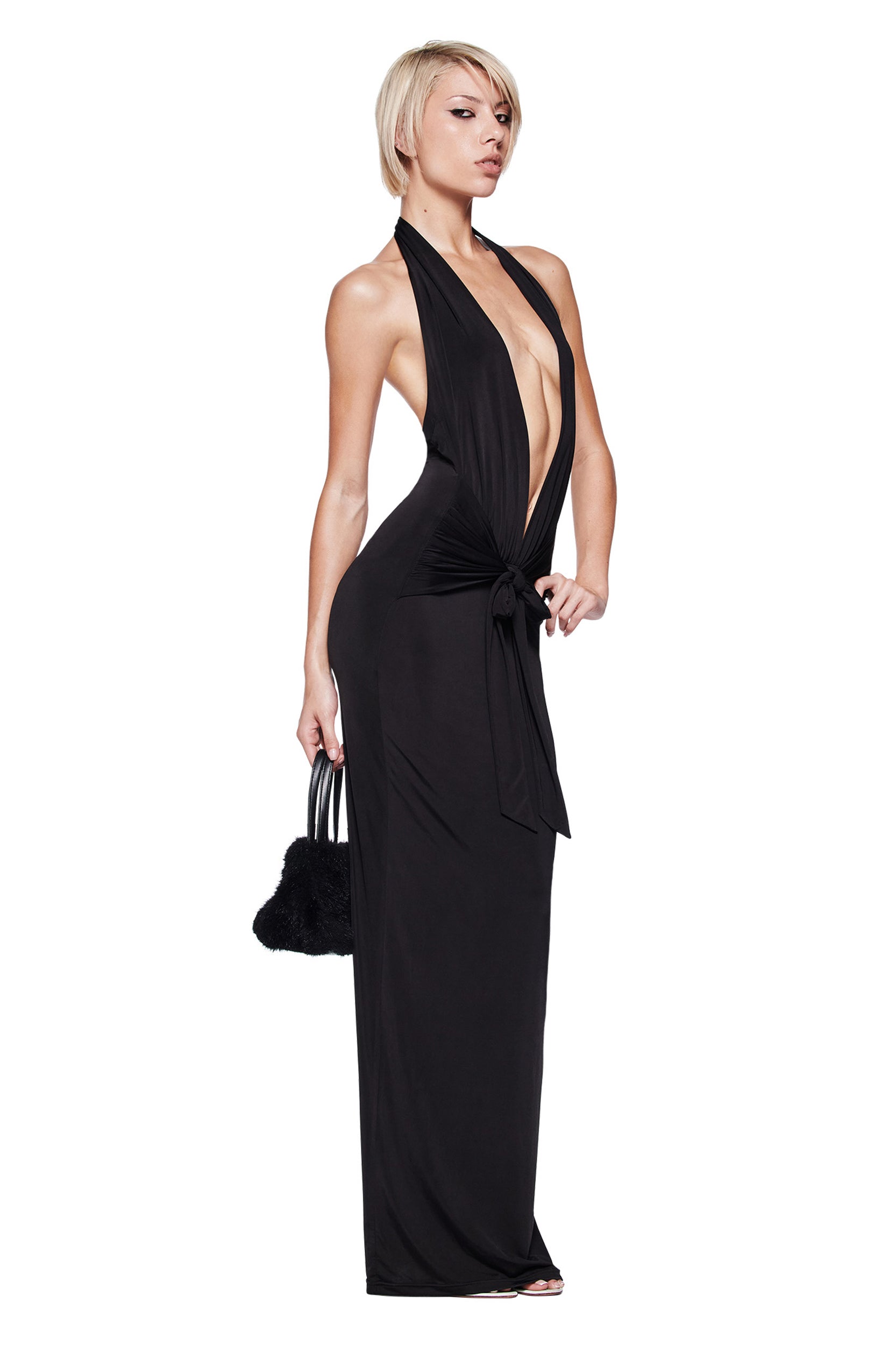 A woman with short blonde hair models the I.AM.GIA ZIVA PLUNGE TIE MAXI DRESS in black, featuring a plunging V neckline and front tie. She holds a small black handbag and poses confidently against a white background.