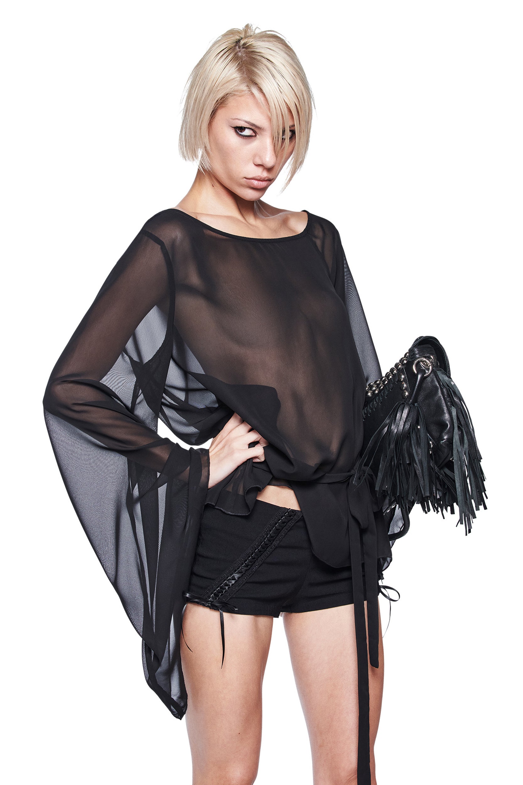 A person with short blonde hair wears the I.AM.GIA MALINA LONG SLEEVE PONCHO TOP in black and black shorts, standing with one hand on their hip and holding a black fringed handbag against a white background.
