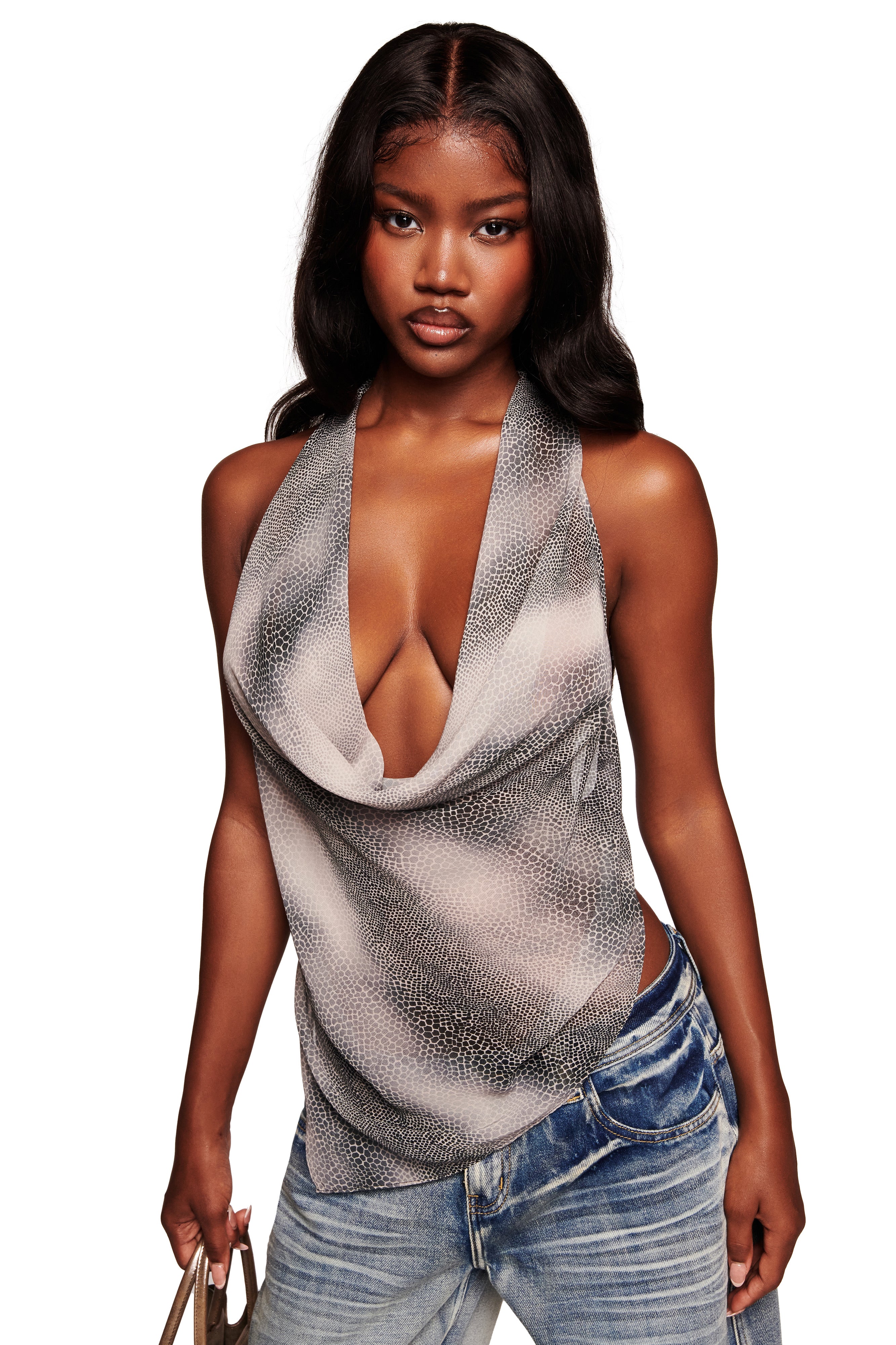 A woman with long, wavy hair wears the I.AM.GIA GRAYSON TOP - SERPICO, a draped sleeveless cowl neck top in gray, paired with faded blue jeans. She holds a small beige handbag and stands against a white background.