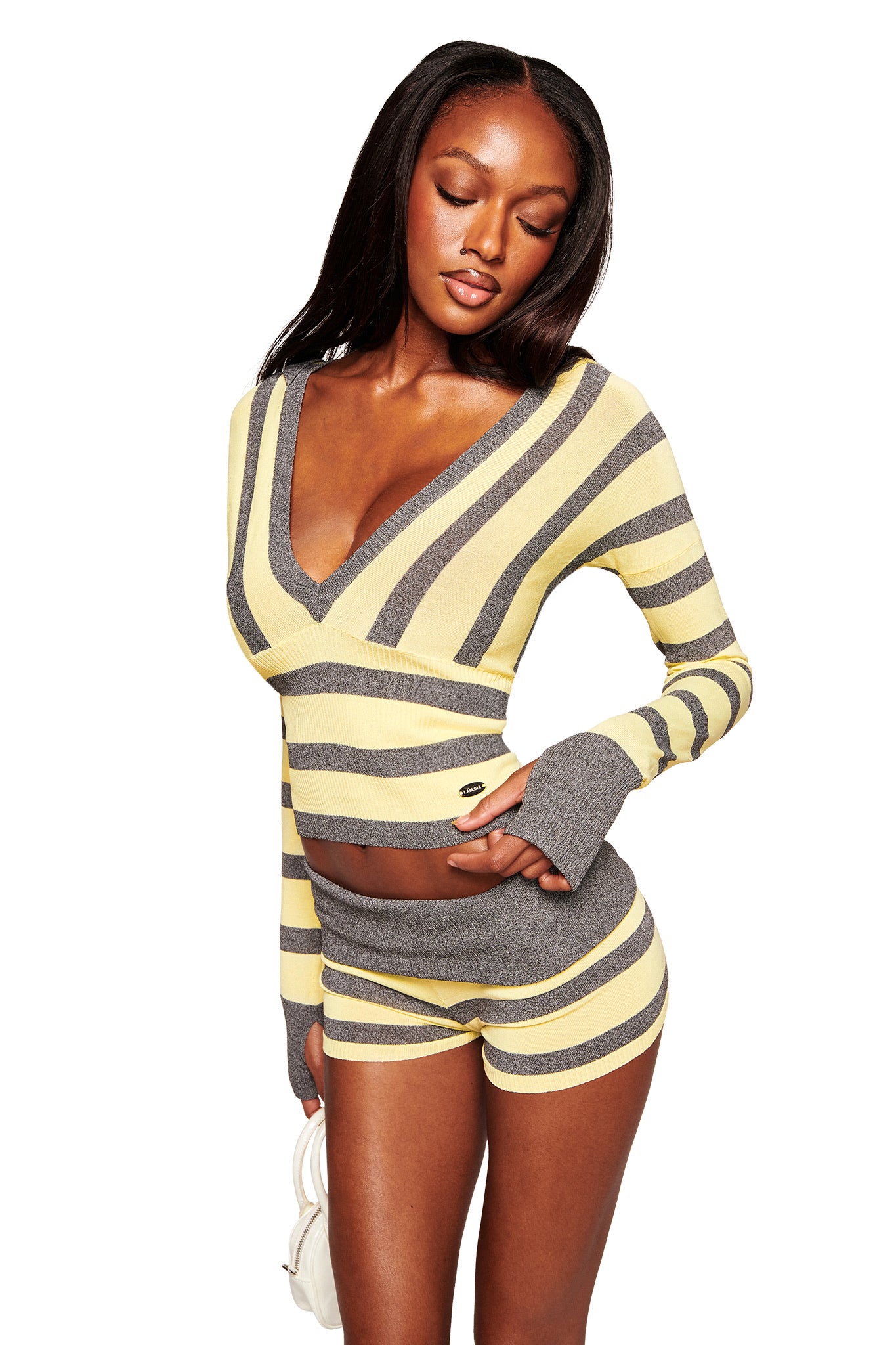 A woman poses in the I.AM.GIA SUZI SWEATER - YELLOW AND GREY STRIPE, a fitted, long-sleeve cropped top with matching high-waisted shorts in soft knit, holding a white purse against a white background.