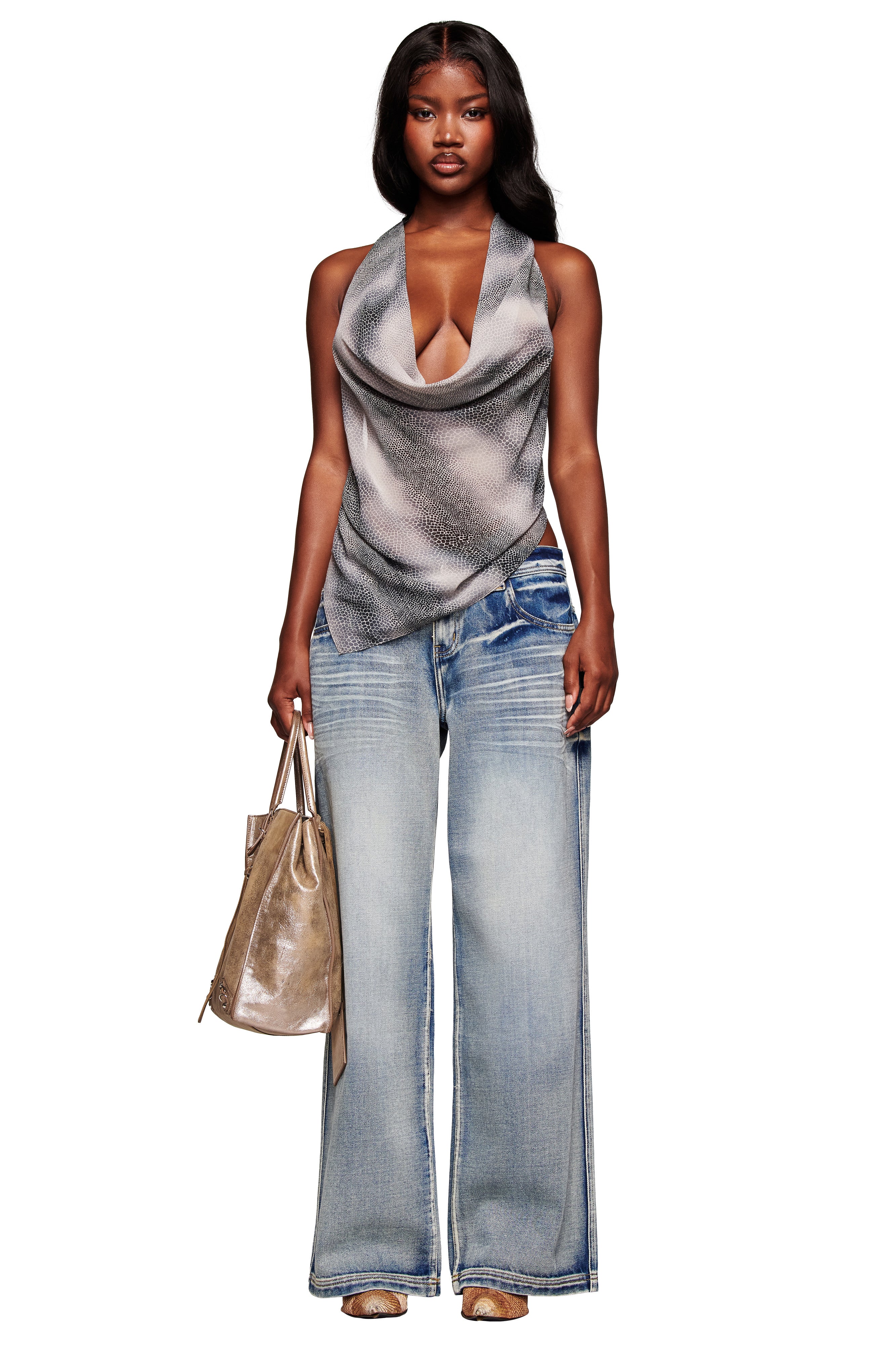 A woman with long dark hair and a confident look holds a metallic tote, wearing the I.AM.GIA PALISADE JEAN - BLUE, a draped gray halter top, tan open-toe heels, and a sleeveless style.