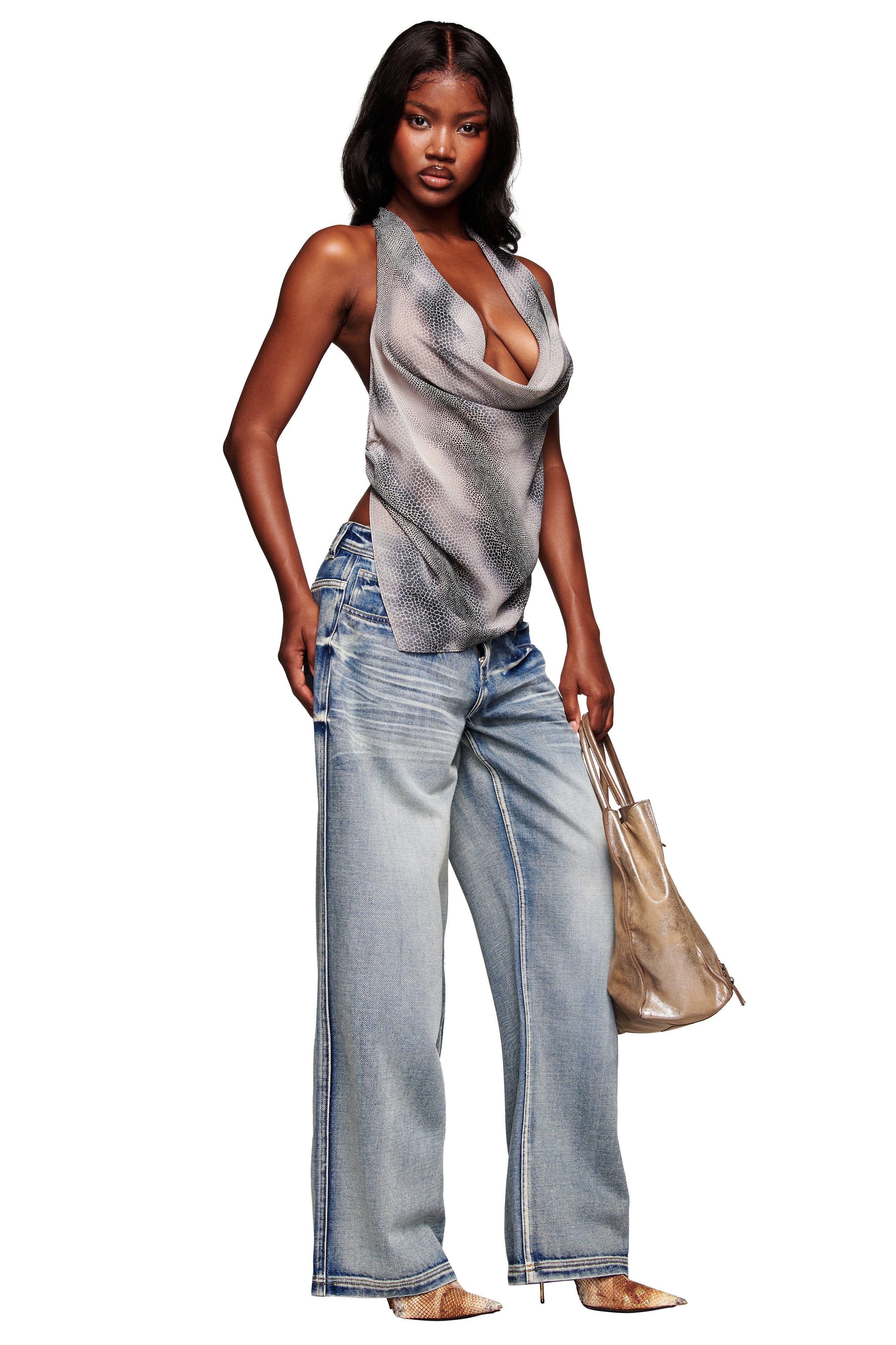 A woman stands confidently in I.AM.GIA's PALISADE JEAN - BLUE, wearing a sleeveless silver draped top, pointed-toe shoes, and holding a metallic gold tote bag.