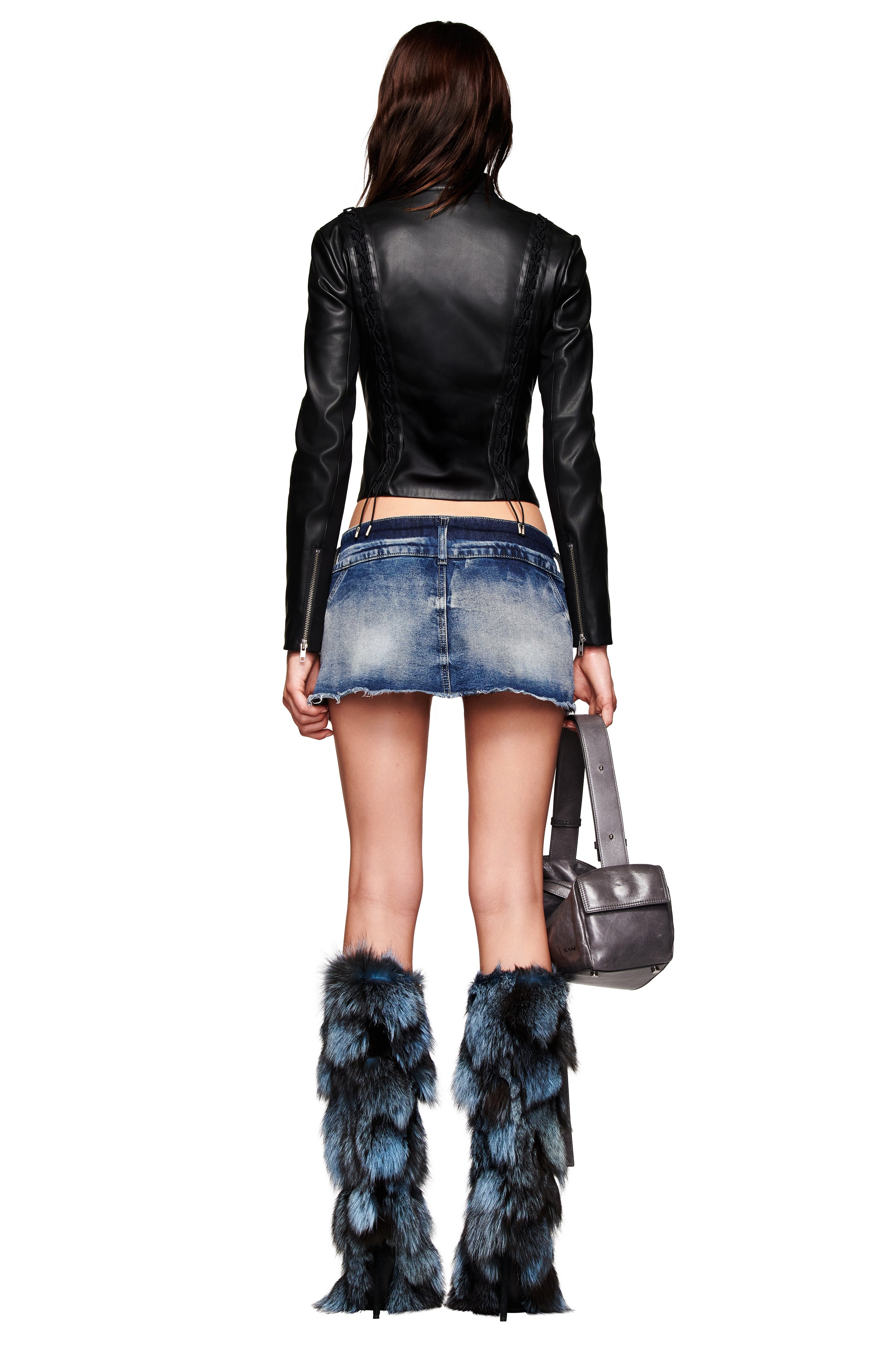 A woman stands facing away, wearing the KENDRIX JACKET - BLACK by I.AM.GIA, paired with a short denim skirt and blue-and-black fur boots, holding a silver handbag.