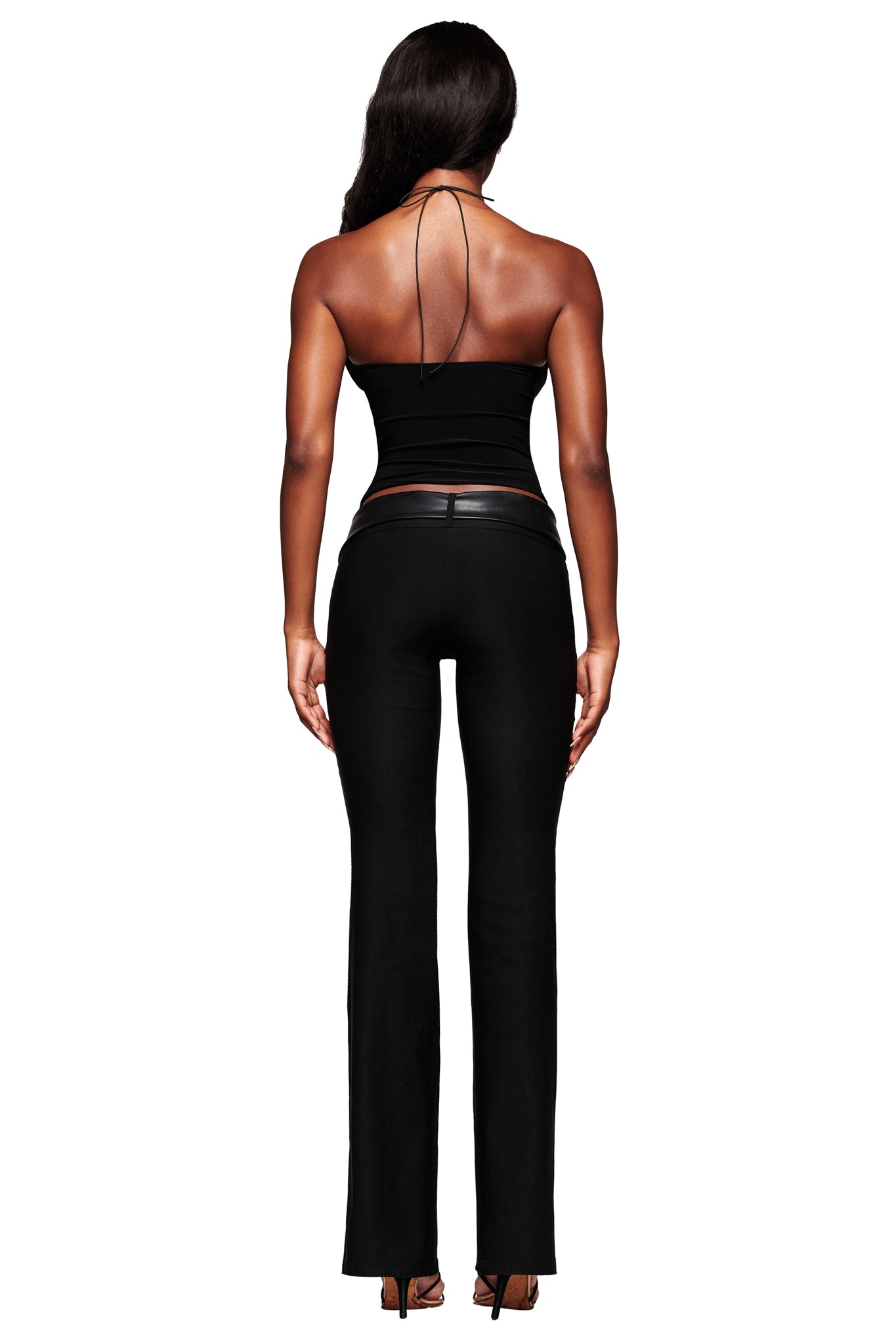 A woman with long dark hair, facing away, wears the I.AM.GIA FENNICK TOP - BLACK with a bodycon fit, styled with fitted black pants and high heels against a white background.