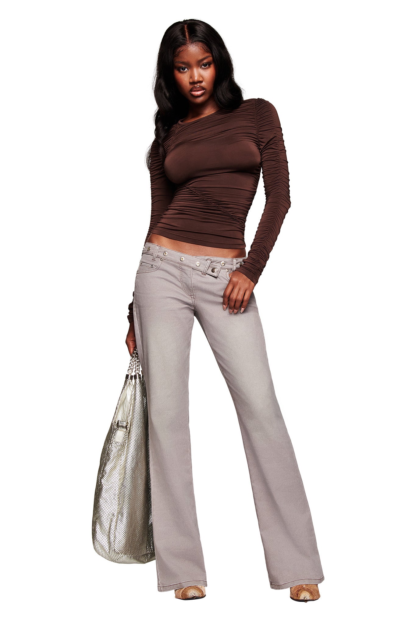 A woman in a fitted brown top and open-toe shoes poses with a large silver handbag, wearing the I.AM.GIA GREER PANT in grey—a light gray wash, low-rise, flared style. Her long dark hair is in loose waves against a white background.