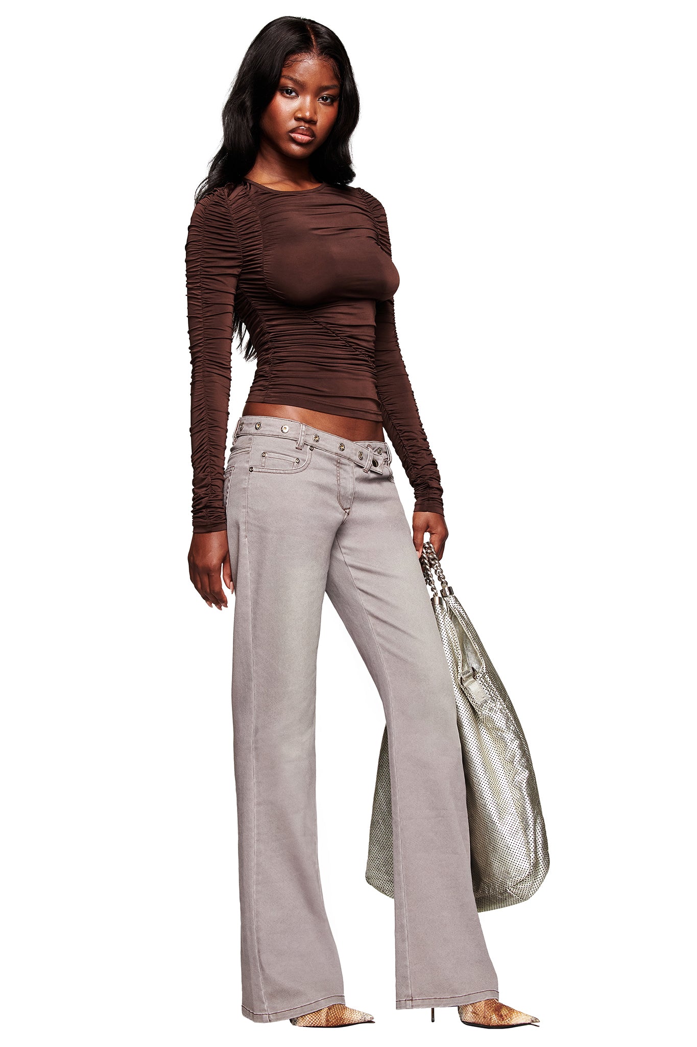 A woman with long dark hair wears the I.AM.GIA GREER PANT in grey, a low-rise, wide-leg style, paired with a fitted brown mesh long-sleeve top, pointed-toe shoes, and carries a large silver tote bag.