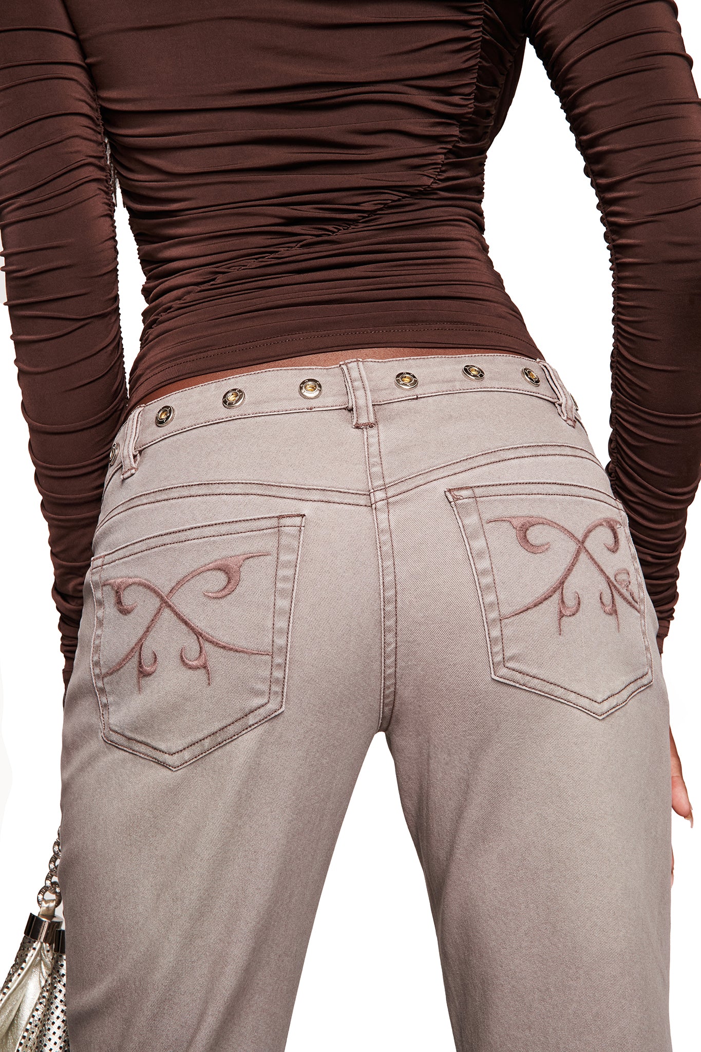 A person, facing away from the camera, wears a ruched brown long-sleeve top and I.AM.GIA GREER PANT - GREY jeans featuring decorative stitching and butterfly embroidery on the back pockets.