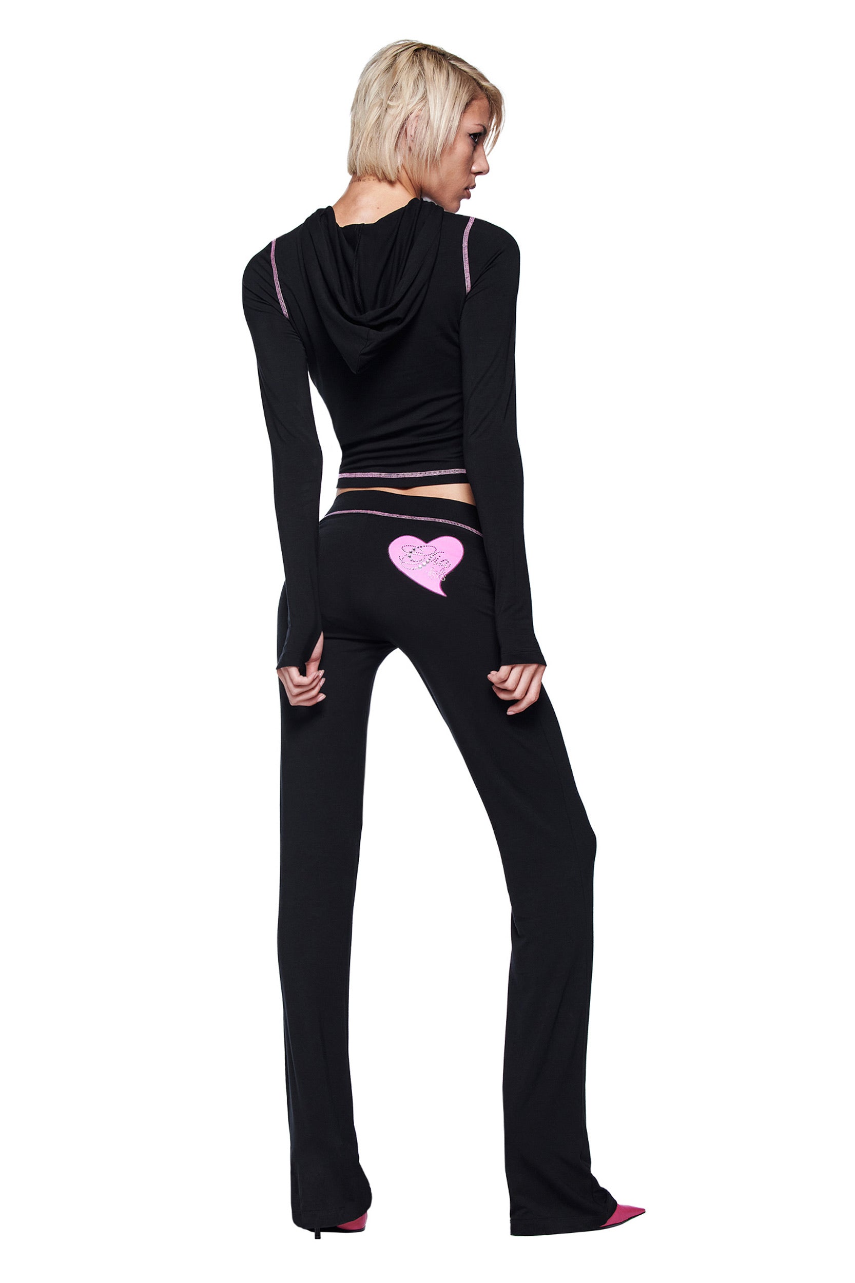 A person with short blonde hair wears I.AM.GIA's Fafi Blare Pant in black, featuring a pink heart on the back, and a black hooded jacket, standing facing away against a white background.