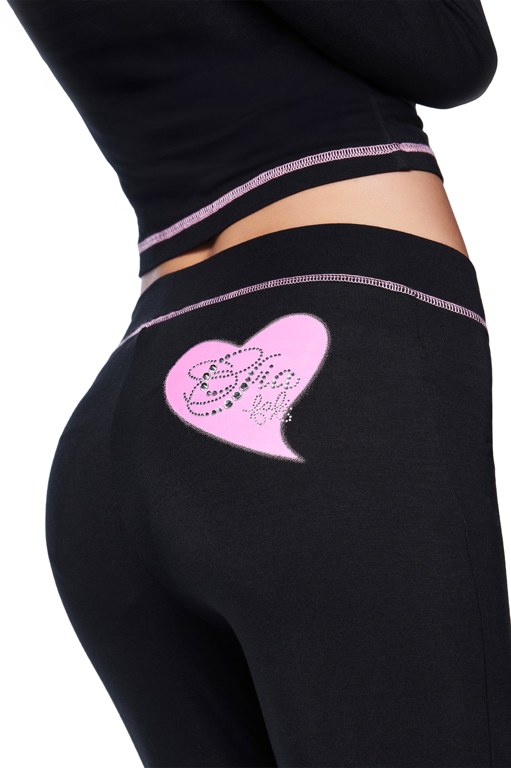 A person wears the I.AM.GIA FAFI BLARE PANT - BLACK and a black top with pink trim; the pants have a pink heart and rhinestone detail on the back.