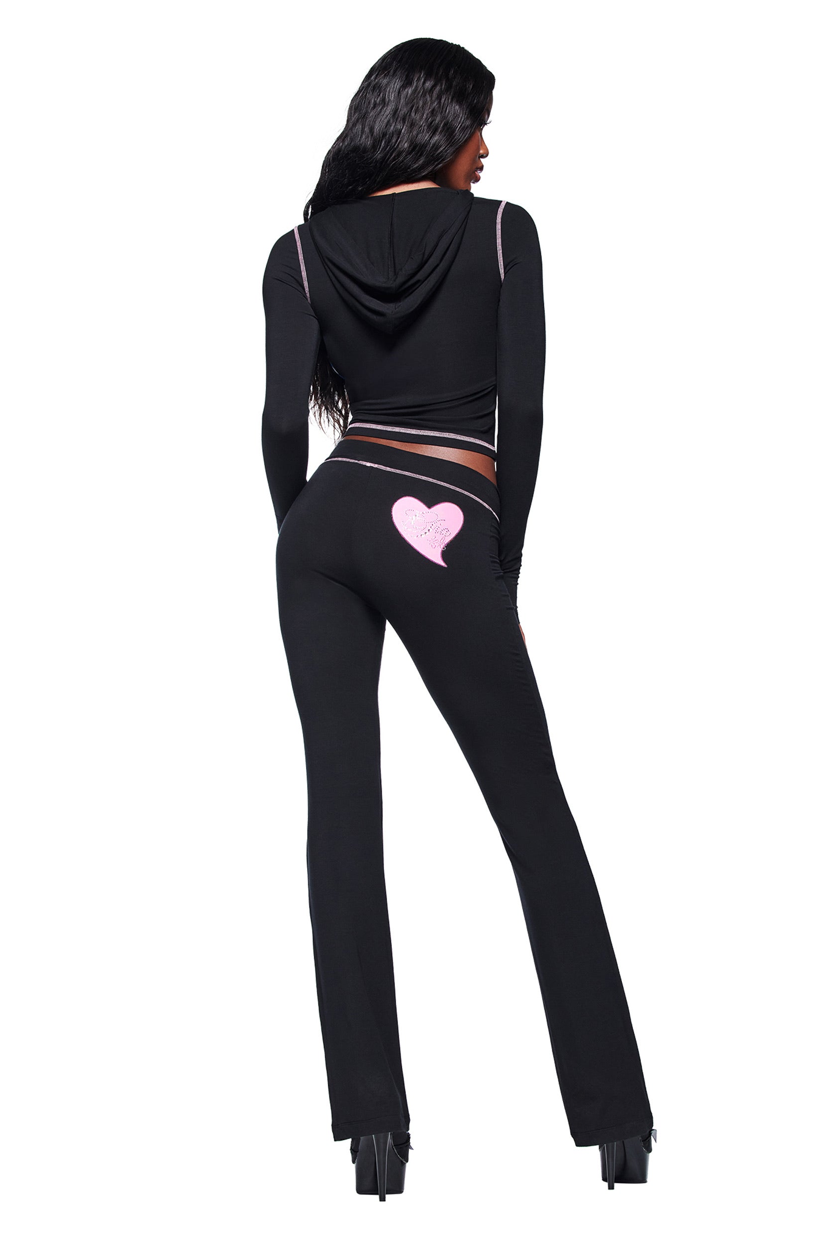 A person stands facing away, hands by their sides, wearing the I.AM.GIA FAFI BLARE HOODIE in black and matching pants with a pink heart design on the back, paired with black platform heels.