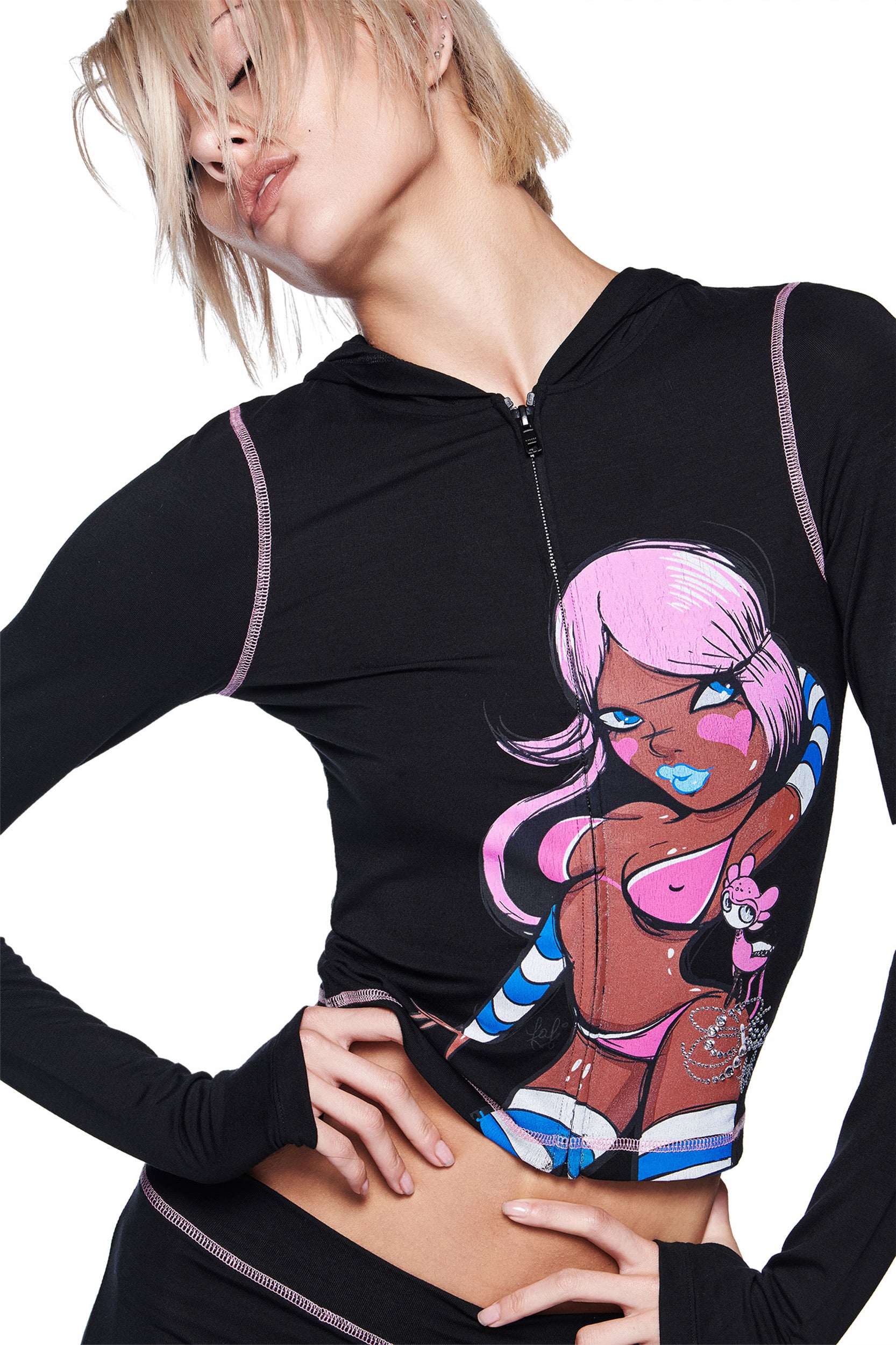 A person with short blonde hair stylishly poses in the I.AM.GIA FAFI BLARE HOODIE - BLACK, which features a colorful cartoon graphic of a pink-haired character in a bikini. One arm is bent, hand resting on their waist.