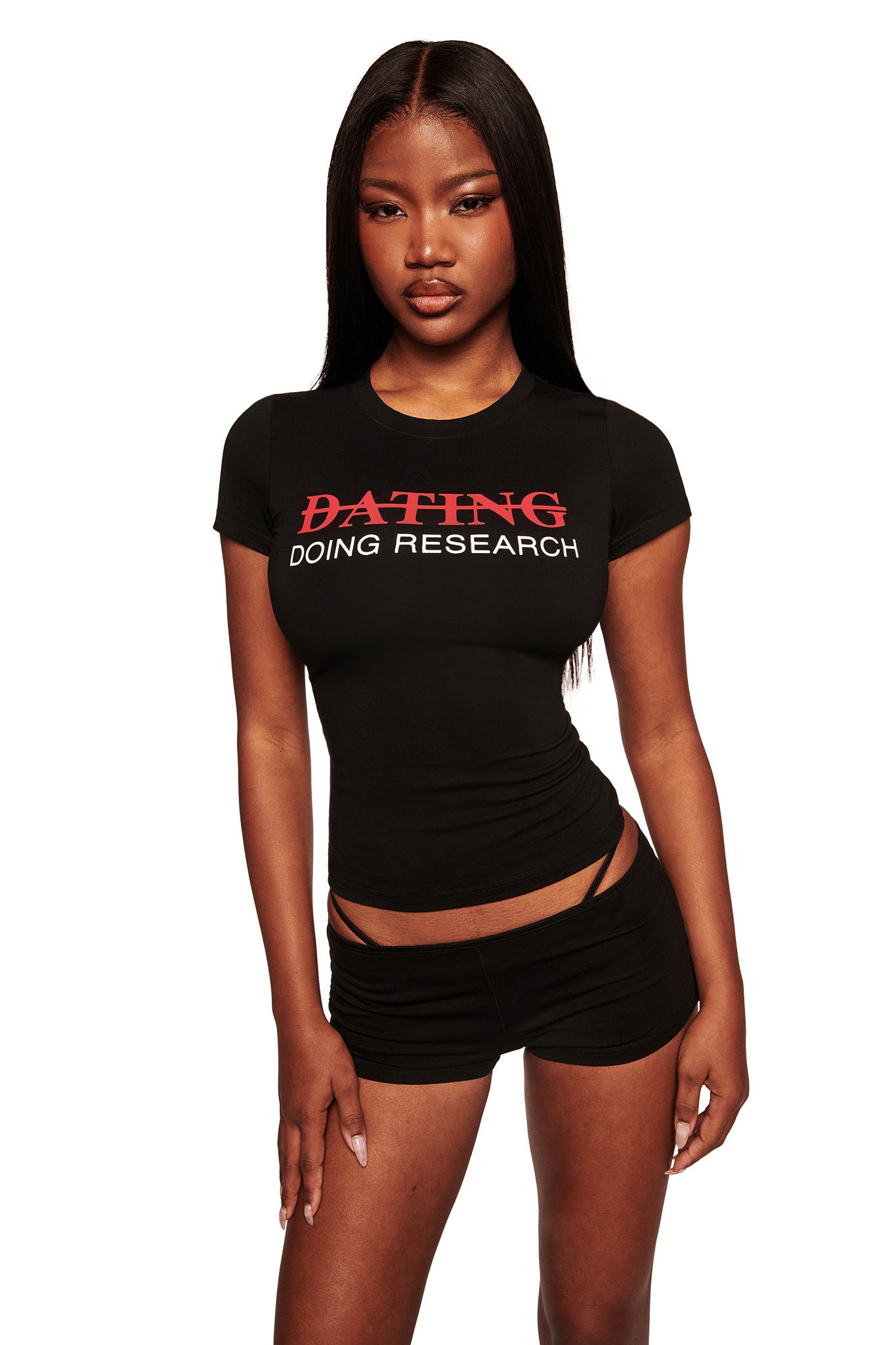 A woman poses against a white background in the POLINA TEE - RESEARCH by I.AM.GIA, a fitted black graphic tee featuring red and white "DATING" (crossed out) and "DOING RESEARCH" text, styled with black shorts.