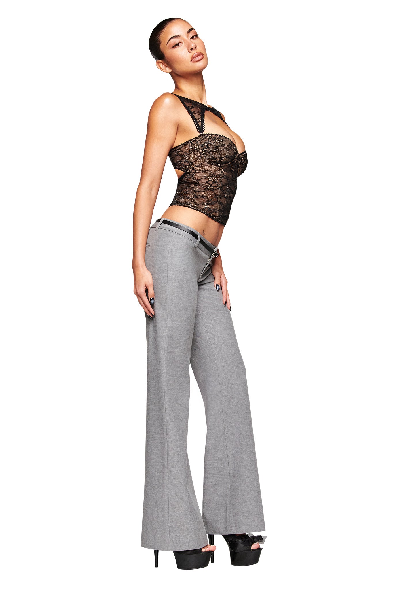 A woman poses confidently in the I.AM.GIA CLARA PANT - GREY, a low-rise, relaxed straight-leg suiting pant, paired with a sheer black lace top and black platform heels against a white background. She looks slightly upward with one hand at her side.