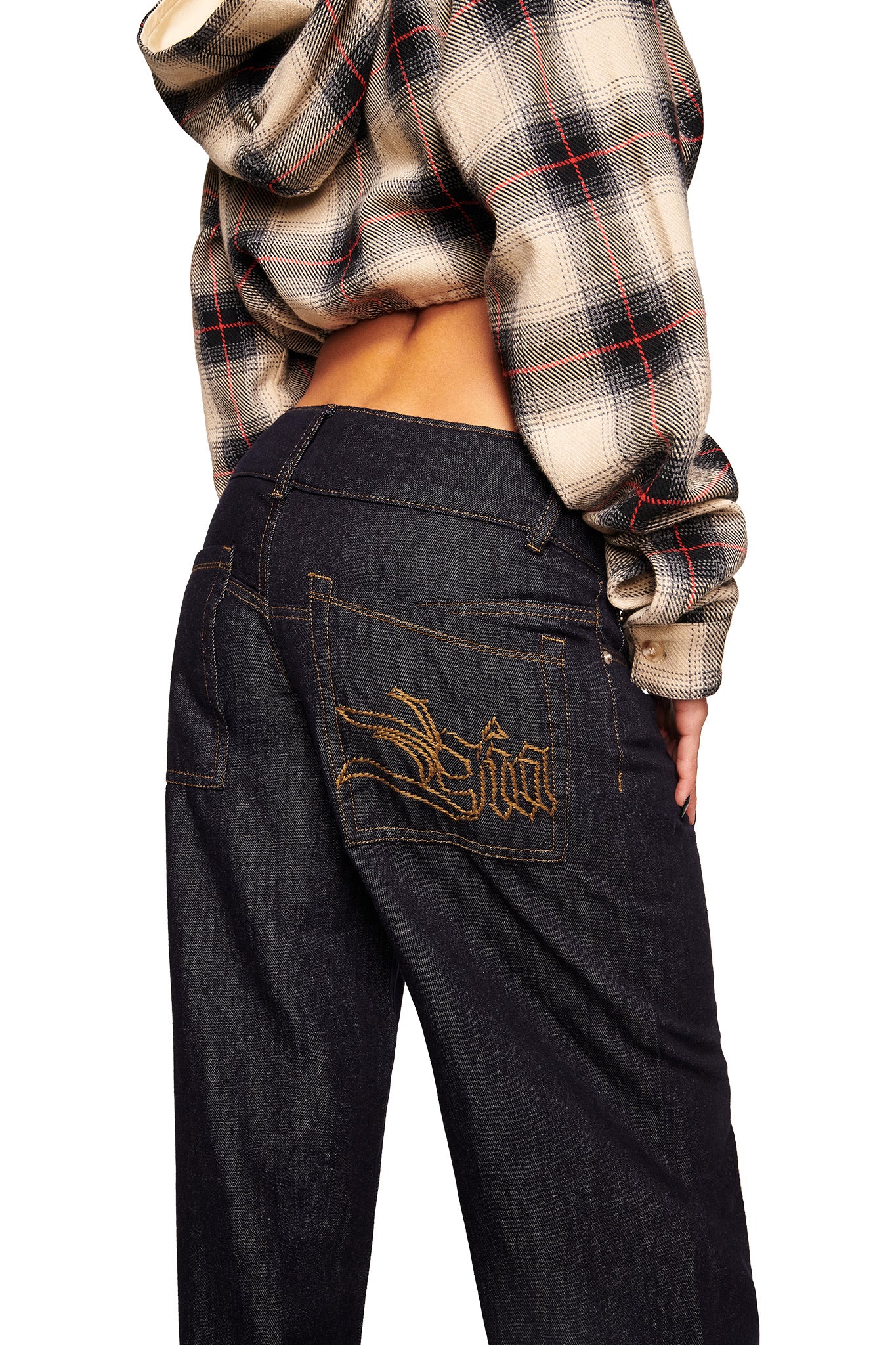 A person wearing a plaid hoodie and I.AM.GIA's MOXIE JEAN - INDIGO stands with their back to the camera. The jeans have a low-rise waist, gold embroidered designs, and writing on the pocket.