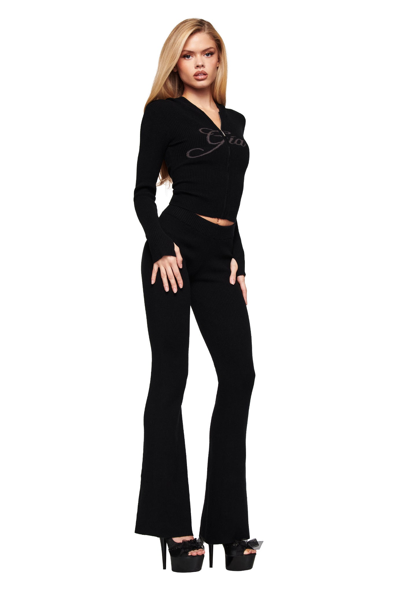 A woman with long blonde hair poses confidently against a white background, wearing the I.AM.GIA BLARE KNIT HOODIE in black paired with matching flared pants and high platform heels.