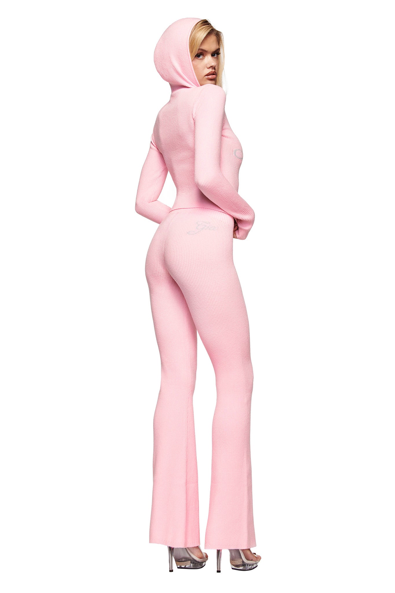 A person models the I.AM.GIA BLARE KNIT HOODIE in pink, featuring a fitted, ribbed design with flared legs, styled with silver heels and posing sideways against a plain white background.