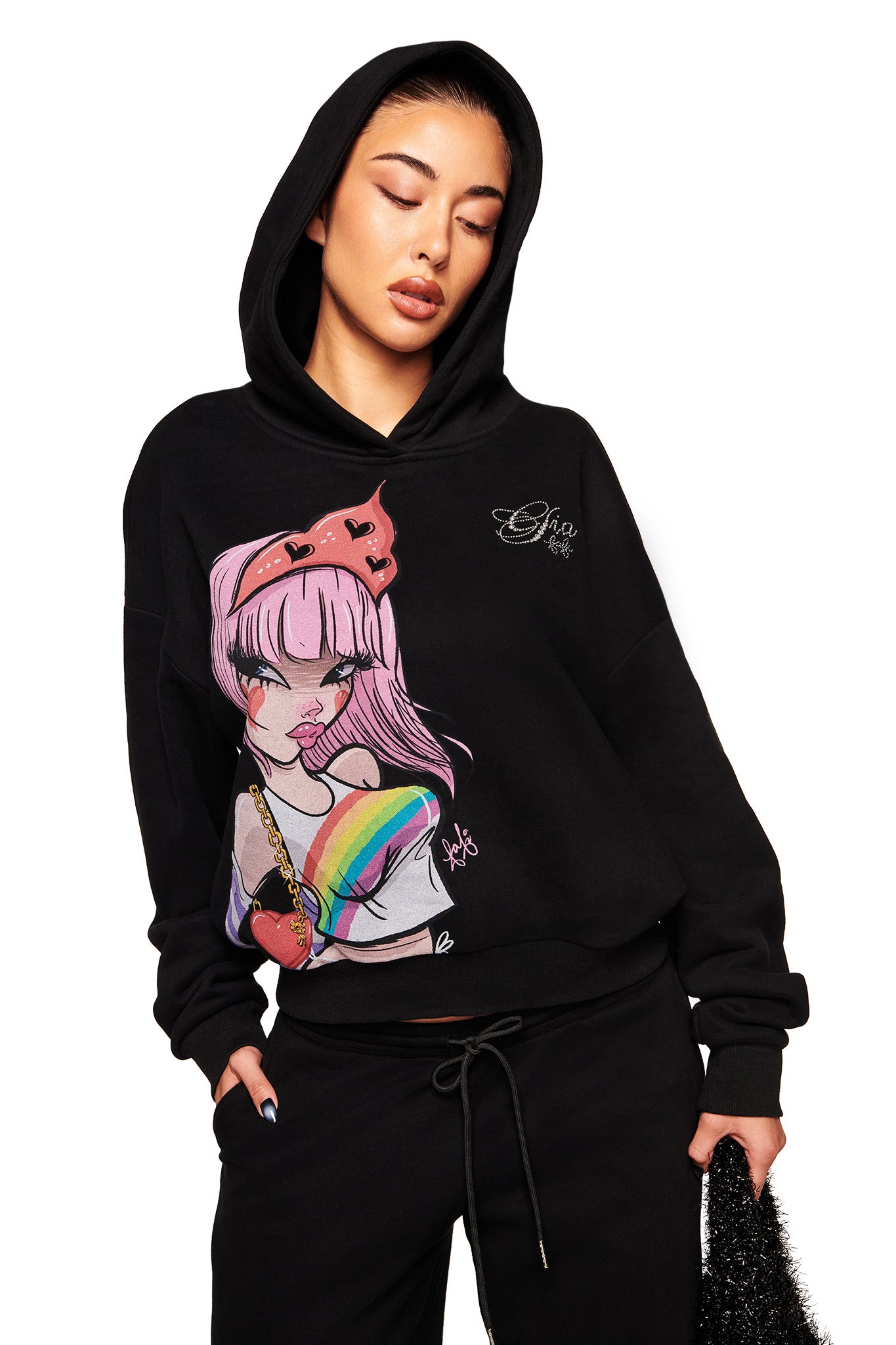 A woman wears the I.AM.GIA FAFI HOODIE - BLACK featuring an anime-style pink-haired girl with a heart and rainbow, part of the FAFI collaboration. Hood up and eyes closed, she styles it with black pants for a relaxed look.