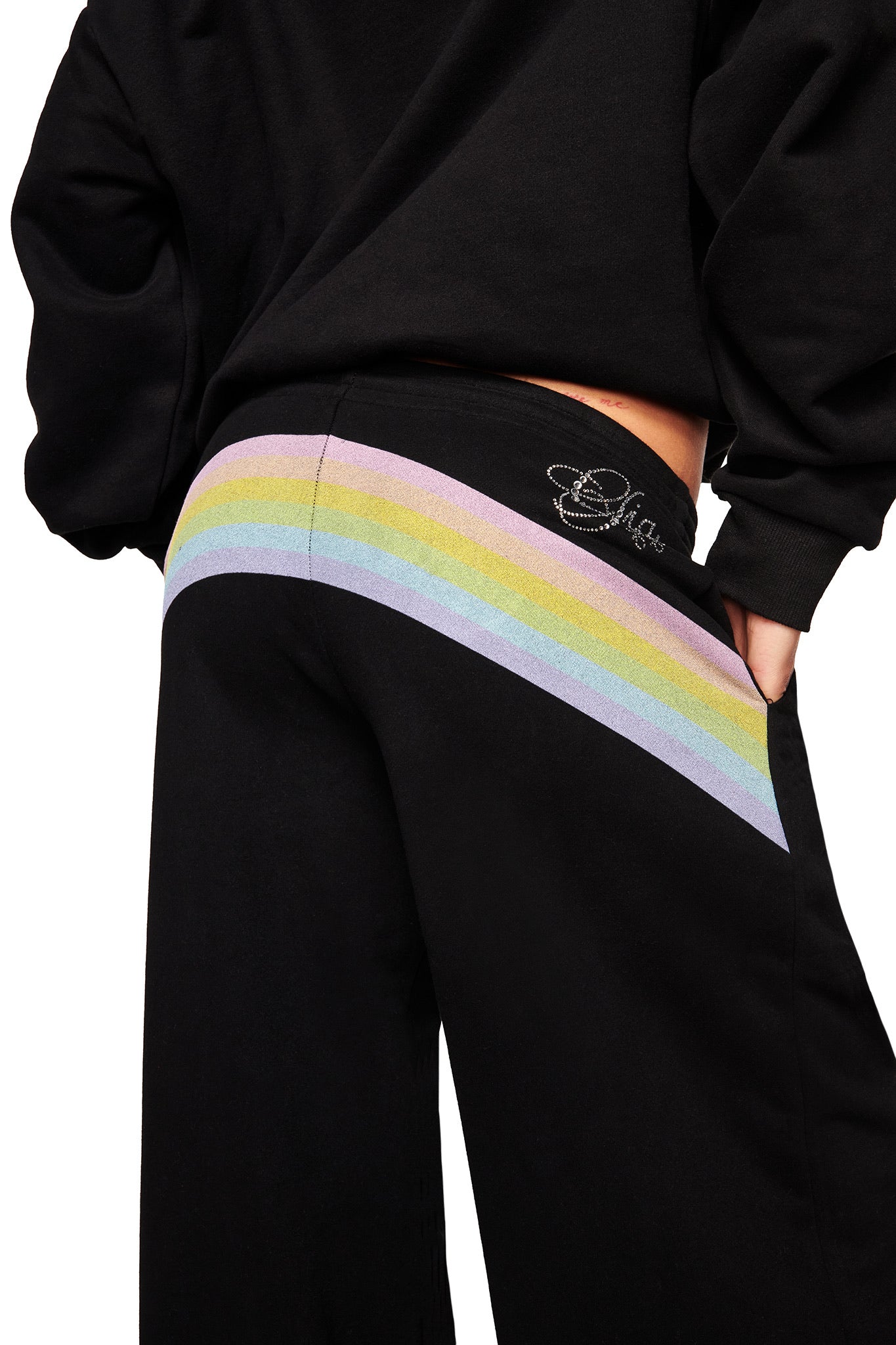 Someone stands with their back slightly turned, wearing the I.AM.GIA FAFI TRACKPANT - BLACK, featuring a pastel rainbow stripe across the back and a sparkly logo on the waistband.